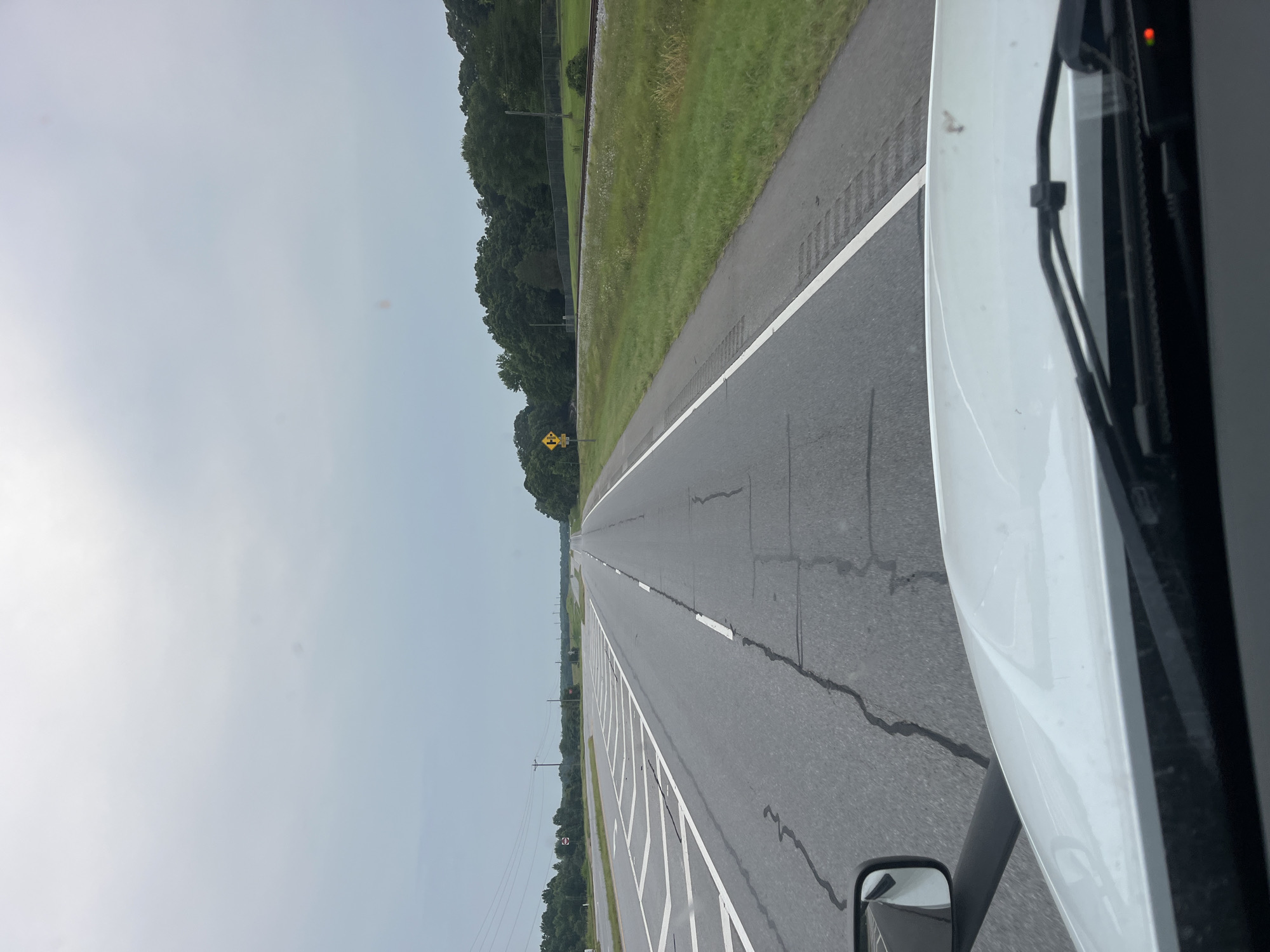 The image captures a view from the cab of a white truck, looking forward on a two-lane highway. The road is asphalt with visible cracks and white lane markings. On the left side of the road, there are complex white markings indicating a merging or diverging lane, and further in the distance, power lines and trees can be seen. To the right of the road, there is a grassy area with a fence and a railroad track running parallel to the highway. A yellow road sign with a black symbol indicating a sharp curve is visible on the right shoulder. The sky is overcast and gray, suggesting a cloudy day. The foreground shows the windshield, wipers, and a side mirror of the truck. The scene appears to be in a rural or suburban setting.