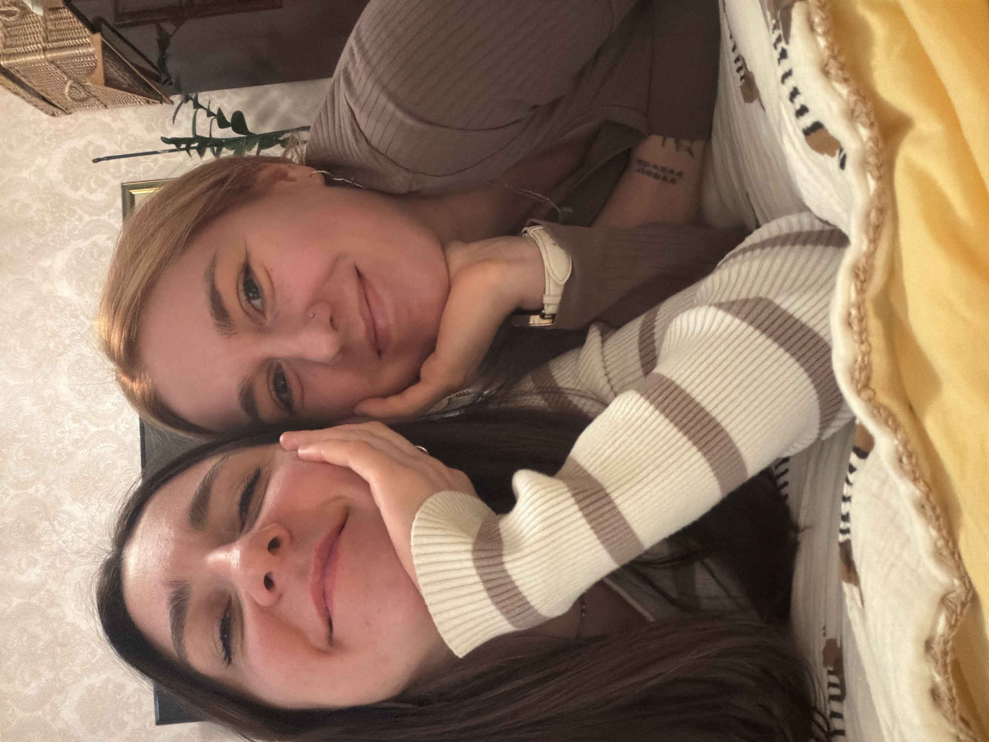 Two young women are depicted in a close-up, sideways-oriented indoor portrait, smiling warmly at the camera. They appear to be lying down, with their heads positioned close to each other, suggesting a moment of intimacy or a shared selfie.
The woman at the top of the frame has light brown hair and a subtle nose piercing. She is smiling with her eyes open and is wearing a ribbed, taupe-colored long-sleeved top. Her left hand is near her chin, and she is wearing a light-colored watch on her left wrist. On her right forearm, a tattoo is visible, featuring Cyrillic script that reads "можна жить и любить," translating to "you can live and love."
The woman at the bottom of the frame has dark hair and is also smiling, with her eyes partially closed, conveying a sense of contentment. She is wearing a cream-colored sweater with horizontal brown stripes. Her hand rests gently near her cheek.
The background features patterned wallpaper with a subtle damask or floral design. Visible behind the top woman are a woven wicker basket, a framed picture on the wall, and a green plant, possibly artificial. A soft yellow blanket or textile is partially visible on the right side of the frame, adding to the cozy indoor setting. The overall mood is one of happiness, affection, and comfort. The Cyrillic text on the tattoo is a notable detail that subtly connects to the Minsk, Belarus location context.