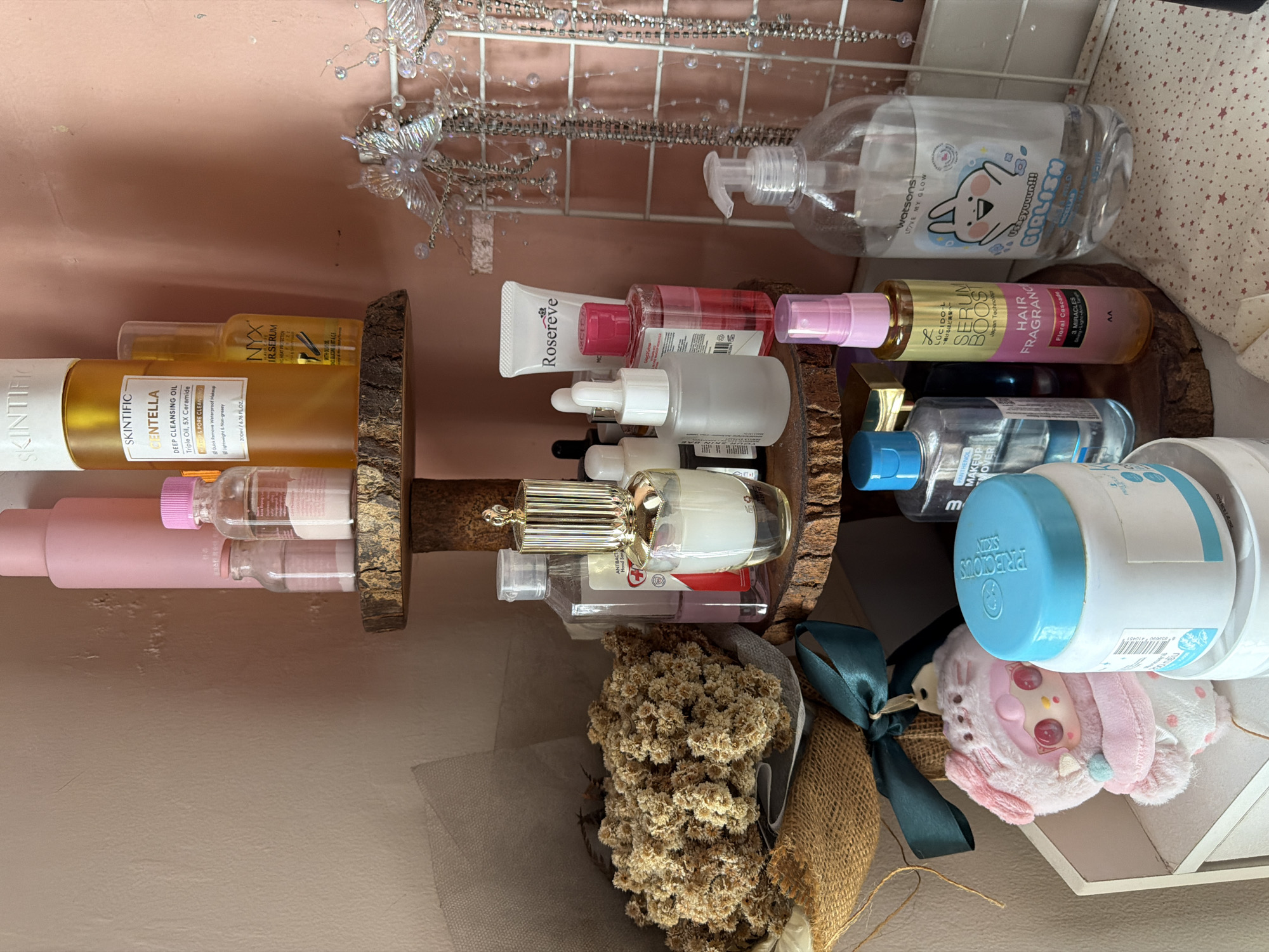 This is an indoor still-life photograph depicting a collection of personal care and cosmetic products arranged on a two-tiered wooden shelf.
The shelf is positioned against a light pink wall. The top shelf contains a bottle of "watsons LOVE MY GLOW" hand sanitizer, a bottle of "LUCIDO-L Serum Boost Hair Fragrance," and a tube of "Rosereve" product, along with several other small bottles.
The bottom shelf holds "SKINTIFIC Centella Deep Cleansing Oil," "NYX Bare With Me Bare With It Serum Concealer," and a variety of other skincare items.
Below the bottom shelf, there is a bouquet of dried flowers wrapped in burlap and tied with a green ribbon. There is also a "Precious Skin" product next to a small plushie toy.
The overall impression is of a personal care product collection displayed in a neat and organized manner. Based on the product labels, this image was likely taken in East Jakarta, Indonesia.