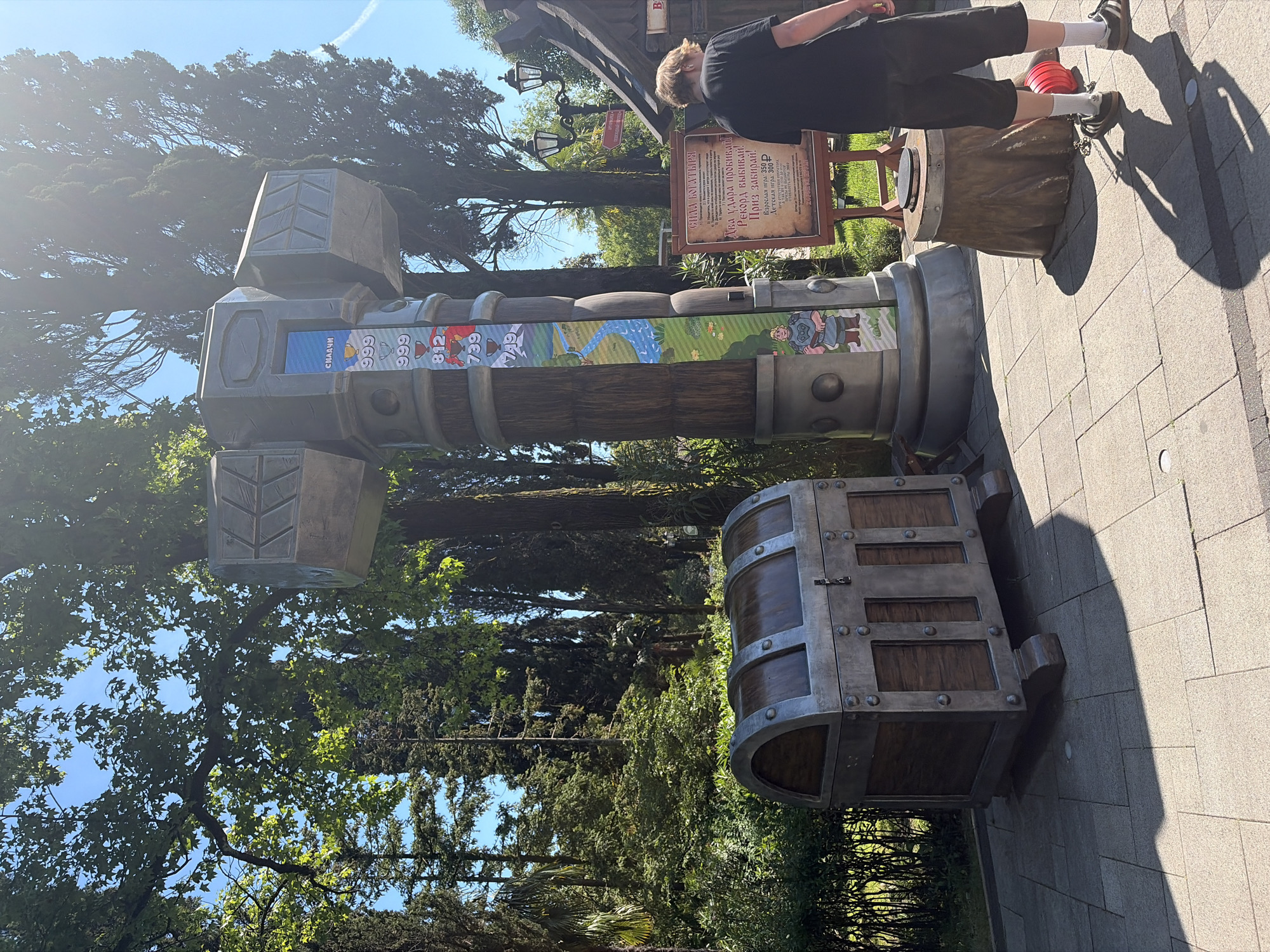 In the city of Sochi, Russia, a person is standing in front of a large, oversized prop hammer, likely part of an attraction in a park or entertainment venue. The hammer has a digital display on its side showing scores or progress, with numbers and trophy icons. Next to the hammer is a wooden chest, also appearing to be a prop. A sign behind the hammer and chest provides information in Russian, possibly detailing rules or costs associated with the attraction. The area is paved, with trees and foliage surrounding the scene, suggesting an outdoor setting. The person, wearing a black t-shirt and shorts, is facing away from the camera, looking towards the attraction. A red object, possibly a bucket or container, is on the ground near their feet. The image is captured in daylight, with shadows indicating a sunny day.