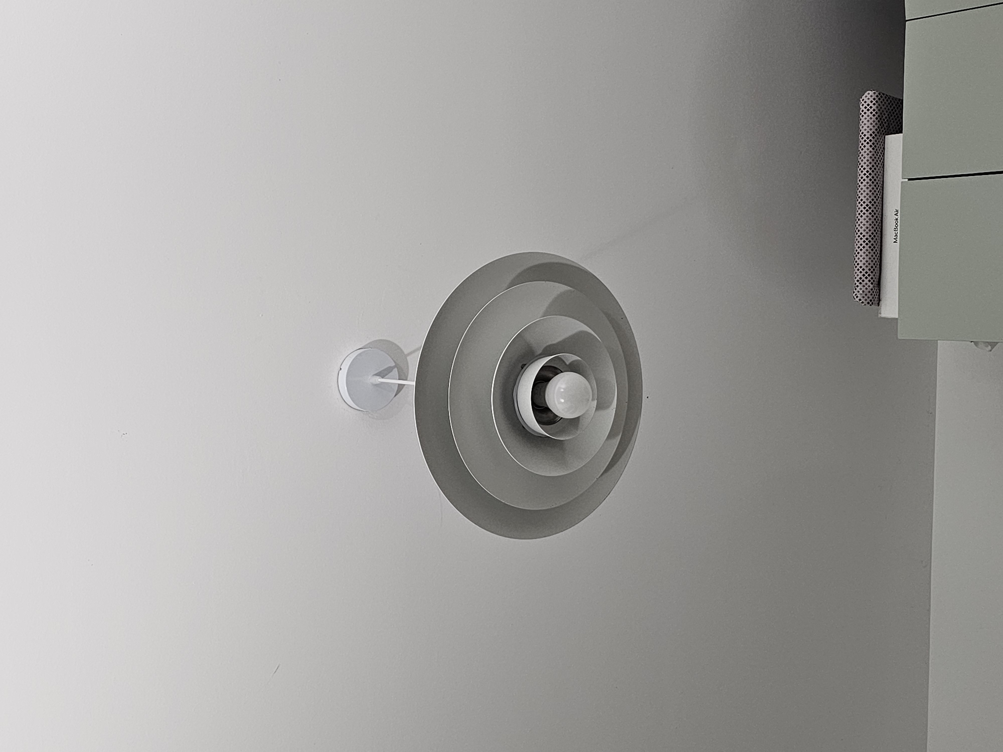 A close-up, low-angle shot shows a pendant light fixture hanging from a white ceiling. The light fixture is made of concentric, matte gray rings that decrease in diameter as they approach the bulb. A single, visible lightbulb is centered within the innermost ring. Below the ceiling, a sliver of a light green cabinet is visible, topped with a white box that appears to be a MacBook Air, and a rolled-up patterned fabric. The image is taken indoors, with no discernible features of a specific city or location.