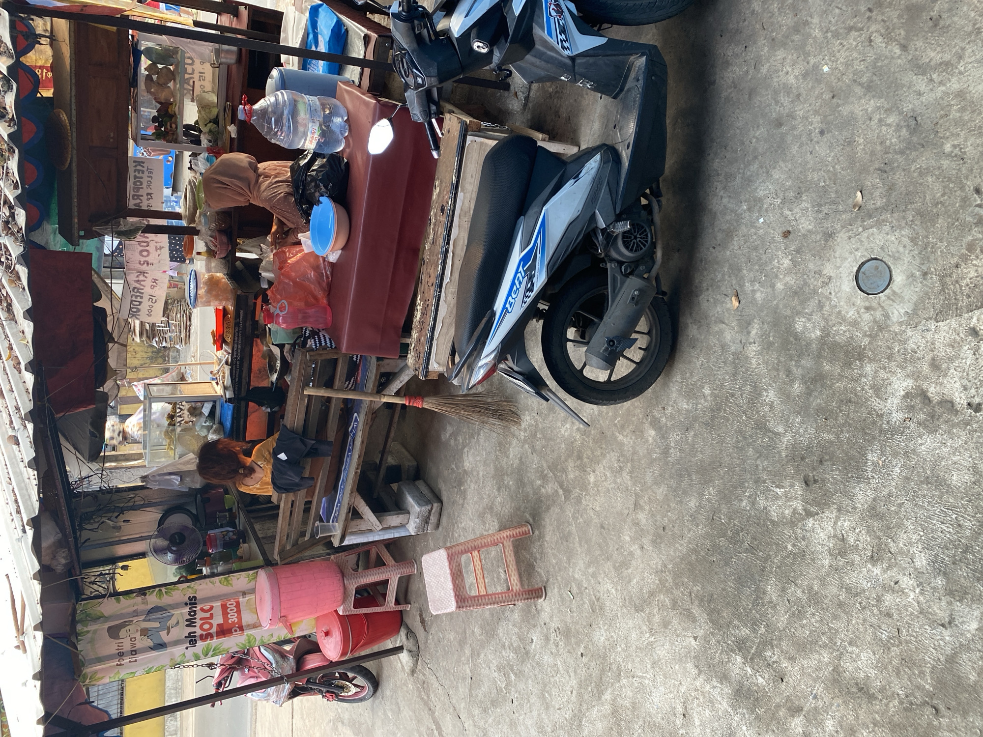 A street food stall in Tangerang, Indonesia, is depicted. A pink motorcycle is parked to the left, and a black and white motorcycle is parked in the foreground to the right. In the center, a woman wearing a headscarf is preparing food. A young girl is seated on a bench behind the food stall. The stall features various food items and drinks displayed on shelves. The ground is made of concrete, and the scene is illuminated by natural daylight.