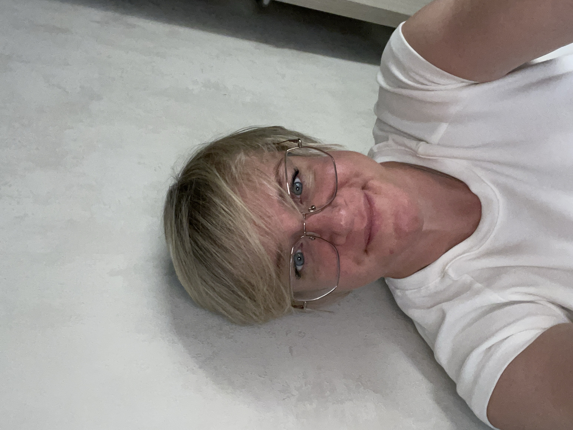 A medium shot shows a woman with short blonde hair and blue eyes wearing glasses and a white t-shirt. She is leaning against a textured, light-colored wall. The woman is smiling and looking at the camera. The background is plain and suggests an indoor setting, possibly a home. There are no specific details to indicate the location in Vologda, Russia.