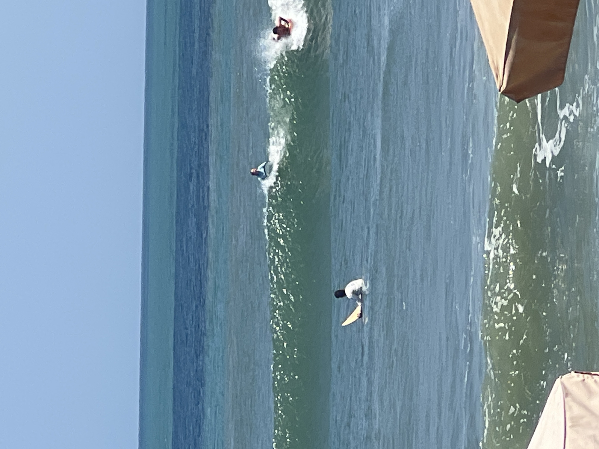 An eye-level, medium shot captures a bright, sunny day at Kuta Beach in Indonesia, with a clear blue sky above a vast expanse of blue-green ocean. Gentle waves are breaking, and several individuals are enjoying the water. In the foreground, partially obscured by a tan beach umbrella, a wave is rolling in. Further out, a person is sitting on a surfboard in the water, facing away from the camera. In the middle distance, a surfer is riding a wave, and another person on a boogie board is also riding a wave. The water is a clear blue-green, with white foam from the breaking waves. The horizon line is distinct where the ocean meets the sky.