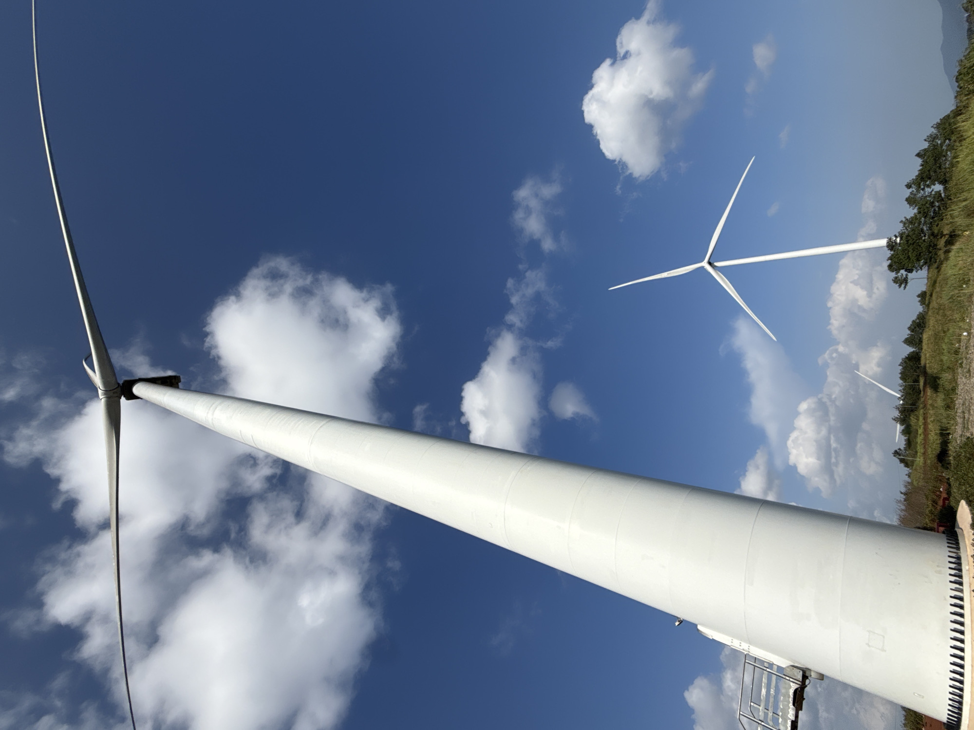 The image captures a low-angle view of a massive, white wind turbine against a bright blue sky with scattered white clouds. The turbine's tall, cylindrical tower dominates the left side of the frame, angled upwards and slightly towards the viewer. A portion of a metal staircase is visible ascending the tower on the left. In the background, another wind turbine stands with its blades extended, silhouetted against the sky. The turbines are situated on a grassy, rolling hillside dotted with trees. The image appears to be taken in a rural or semi-rural setting, possibly part of a wind farm. While the image is described as being in Trường Thọ, Vietnam, there are no specific landmarks or signs visible in the image to confirm this location. The scene is static, with no people or animals present. The lighting suggests it is daytime, likely under sunny conditions.