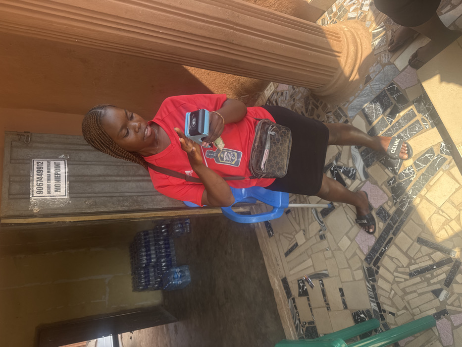 The image depicts a young adult female standing on an irregularly tiled outdoor or semi-outdoor surface in Aguata, Nigeria. The subject has dark skin and braided hair, wearing a red short-sleeved t-shirt with a dark bottle logo, a black skirt, and black "Celine" branded slide sandals. A dark brown patterned crossbody bag is visible on her left side, and a gold-colored bracelet is on her right wrist. She holds a light blue and black portable point-of-sale (POS) device in her left hand and is operating its screen with her right index finger. Nigerian Naira banknotes are partially visible in her left hand beneath the device.
In the background, a metallic door features a white sign displaying "9067449912", "OKORO CHIKA MARYANN", and "MONIEPOINT". To the left of the door, an open doorway reveals an interior area with pale yellow walls, where multiple stacks of shrink-wrapped plastic water bottles are visible on the floor. A blue plastic chair is partially obscured behind the subject. To the subject's right, a fluted brown pillar is prominent, casting a shadow due to strong natural sunlight. The ground surface is composed of a mosaic of light, dark brown, and black tile fragments. The lower right foreground shows the partial view of another individual's legs and feet, wearing brown sandals.