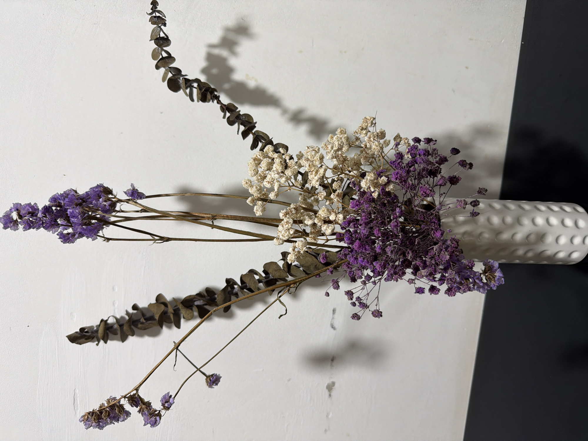 A dried floral arrangement, featuring purple statice, white baby's breath, and brown eucalyptus leaves, is displayed in a white, textured vase. The vase sits on a black surface, casting a shadow against a white wall. The lighting suggests an indoor setting, possibly a home or an office space. The image does not contain any information that can be used to determine its location within Davao City, Philippines, or any other specific geographic location.