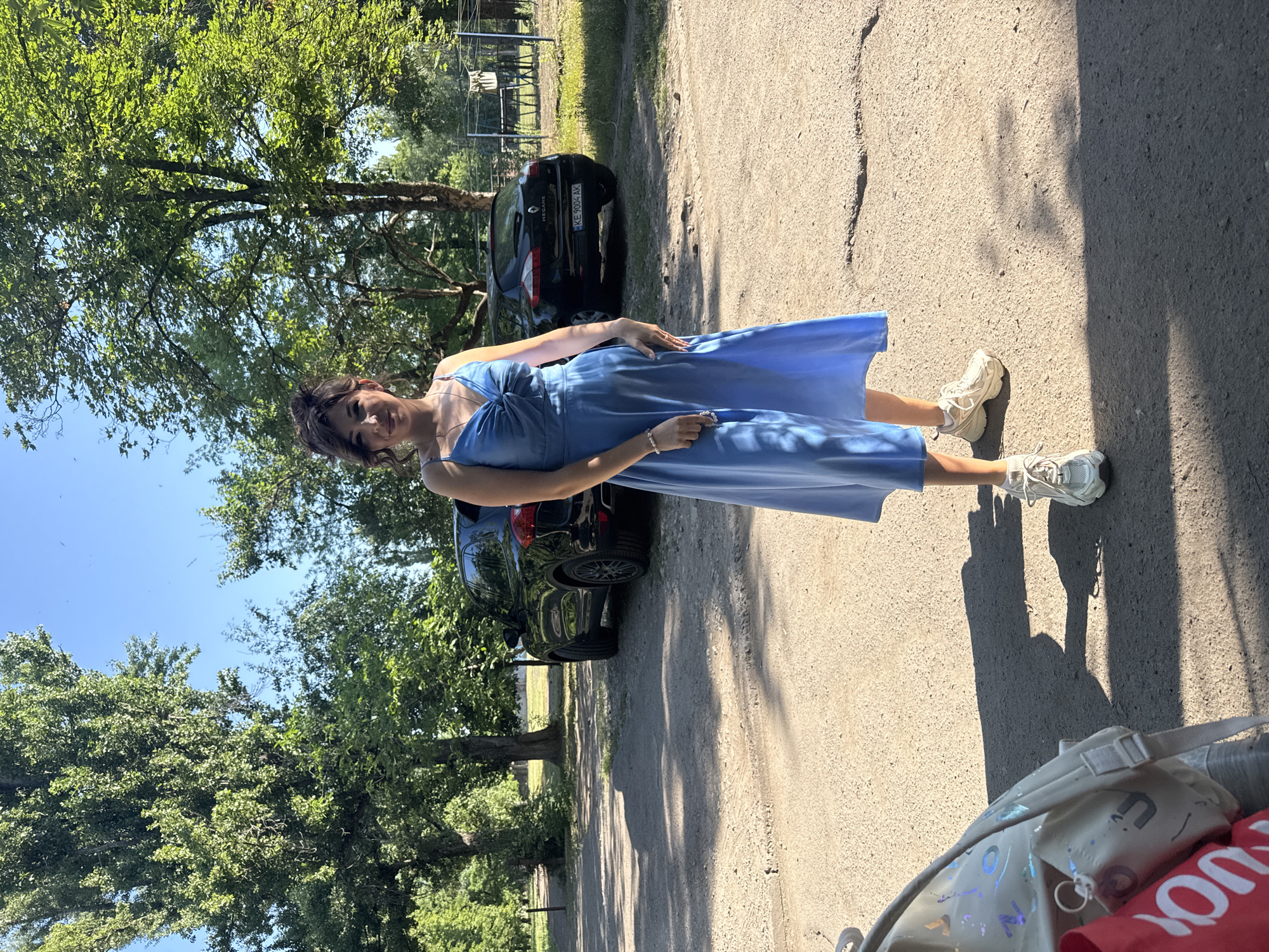 A young woman stands on a dirt road in a park-like setting on a sunny day. She is wearing a blue sleeveless dress with a twisted neckline and white sneakers. Her hair is dark and styled in an updo. She is looking towards the camera with a slight smile. In the background, there are trees and two dark-colored cars parked on the side of the road. A bag with a red component is visible in the lower left corner of the frame. The scene appears to be outdoors in a somewhat rural or suburban area. The plate on one of the cars reads "KE 9004 AK".