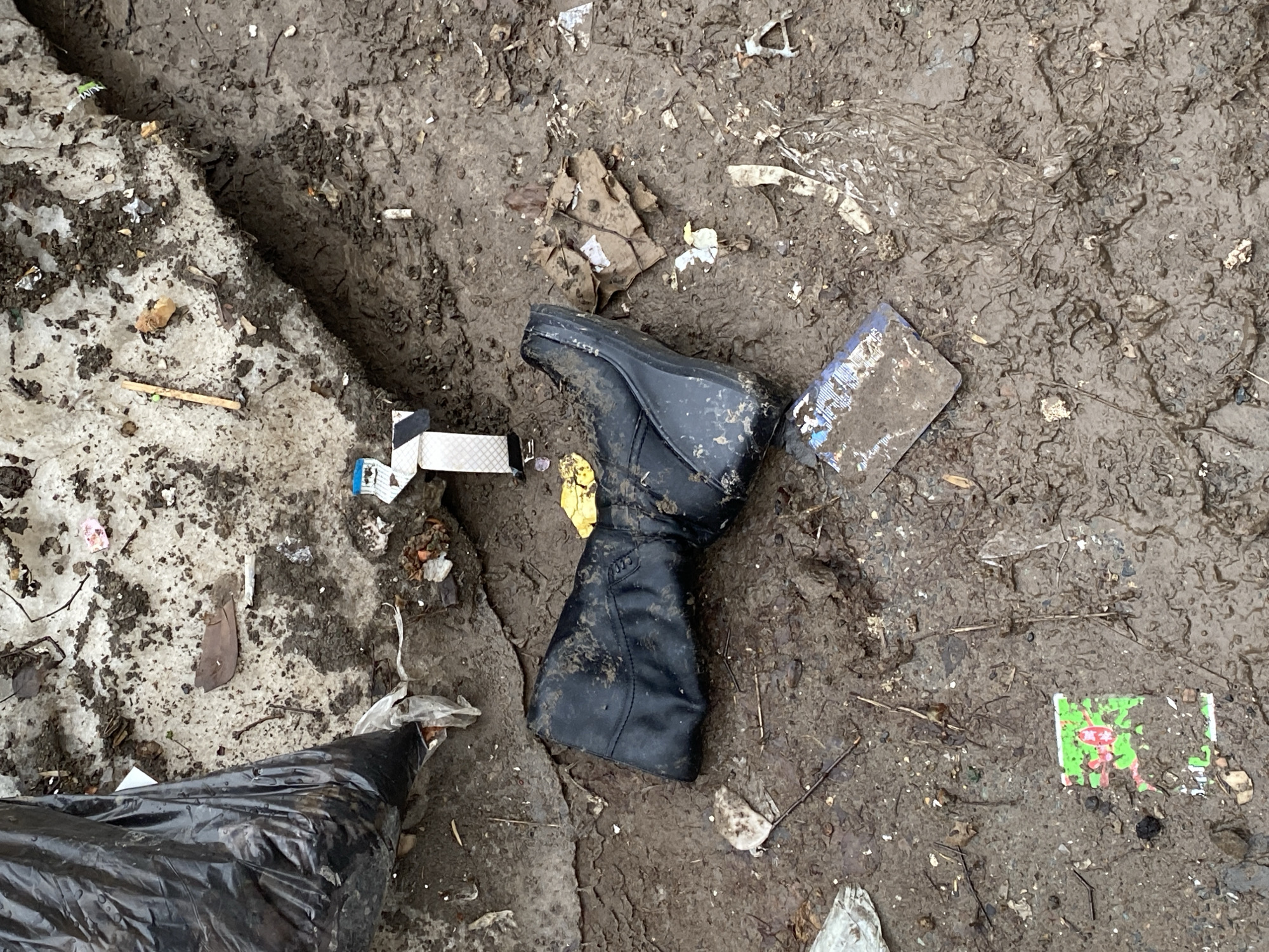 The image depicts a muddy area littered with trash, possibly in Votkinsk, Russia. The ground is covered in mud, small pieces of litter, and debris. A black boot lies horizontally in the center of the scene, partially embedded in the mud. Near the boot are a few rectangular objects appearing to be discarded packaging or containers. The edges of the scene show a mix of melting snow and more accumulated trash. A black plastic bag is visible in the lower left corner of the frame.