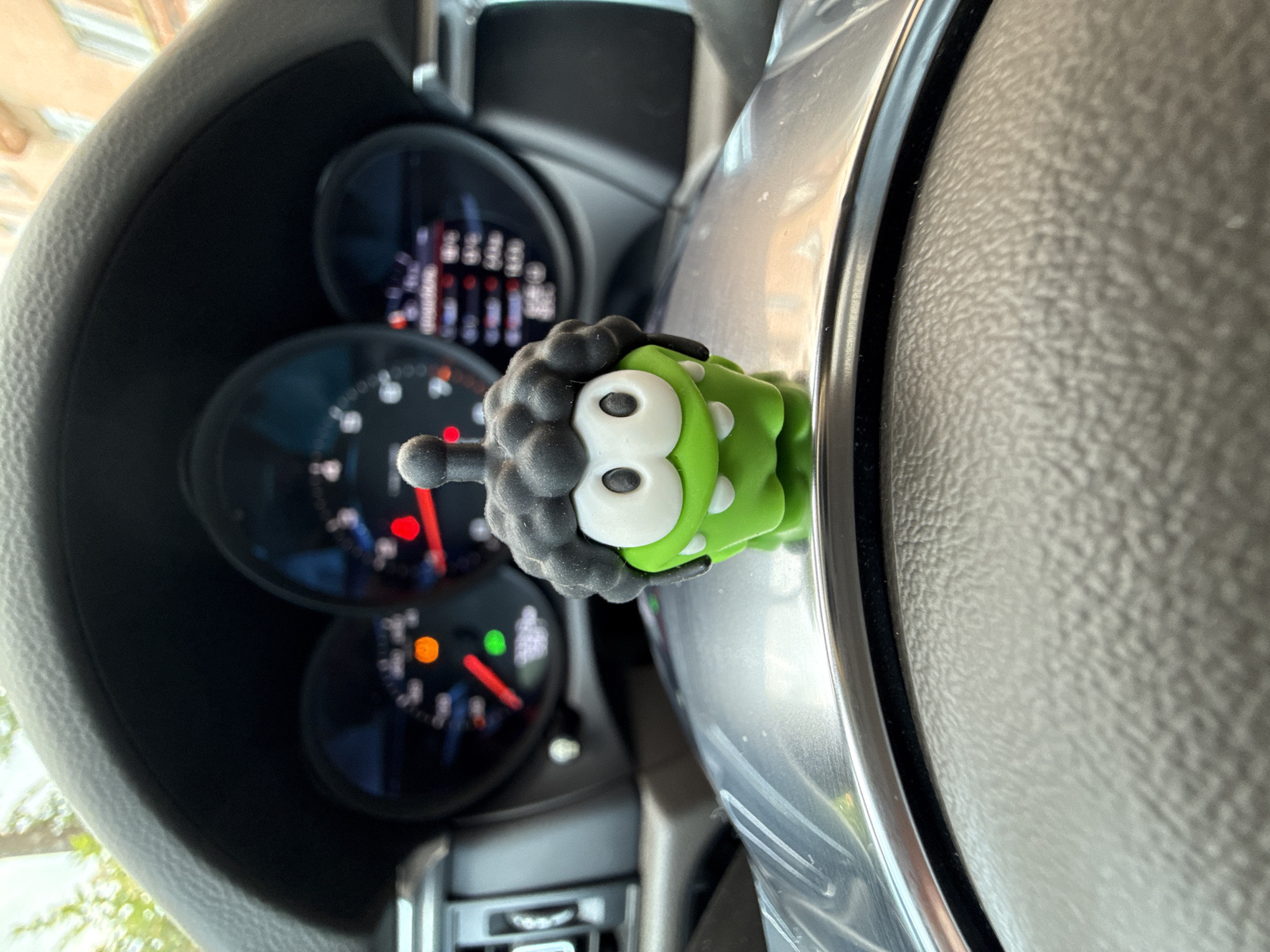A close-up shot taken inside a car. A green and black Om Nom toy with large white eyes is attached to the car's steering wheel. The dashboard gauges are visible behind the toy.