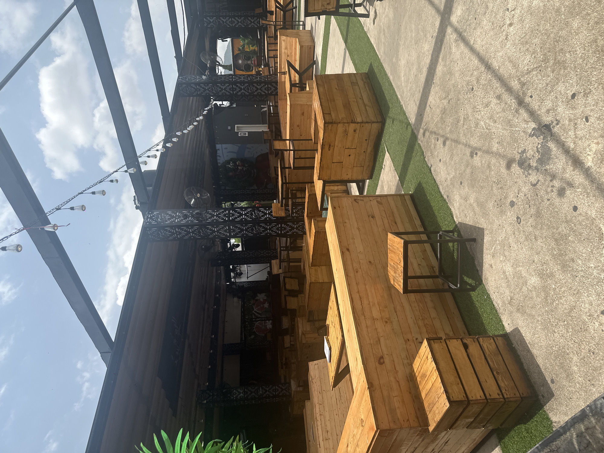 The image depicts an outdoor seating area of a commercial establishment, likely a restaurant or bar, situated in Howayi, Nigeria. The area is covered by a dark metal pergola with exposed beams and a corrugated roof. String lights are suspended from the pergola. The seating consists of modular wooden benches and tables, with some areas featuring artificial turf interspersed with concrete flooring. In the background, the establishment appears to have a darkened interior with some artwork visible on the walls. There are no people visible in the image. The overall impression is a relaxed, open-air dining or gathering space.