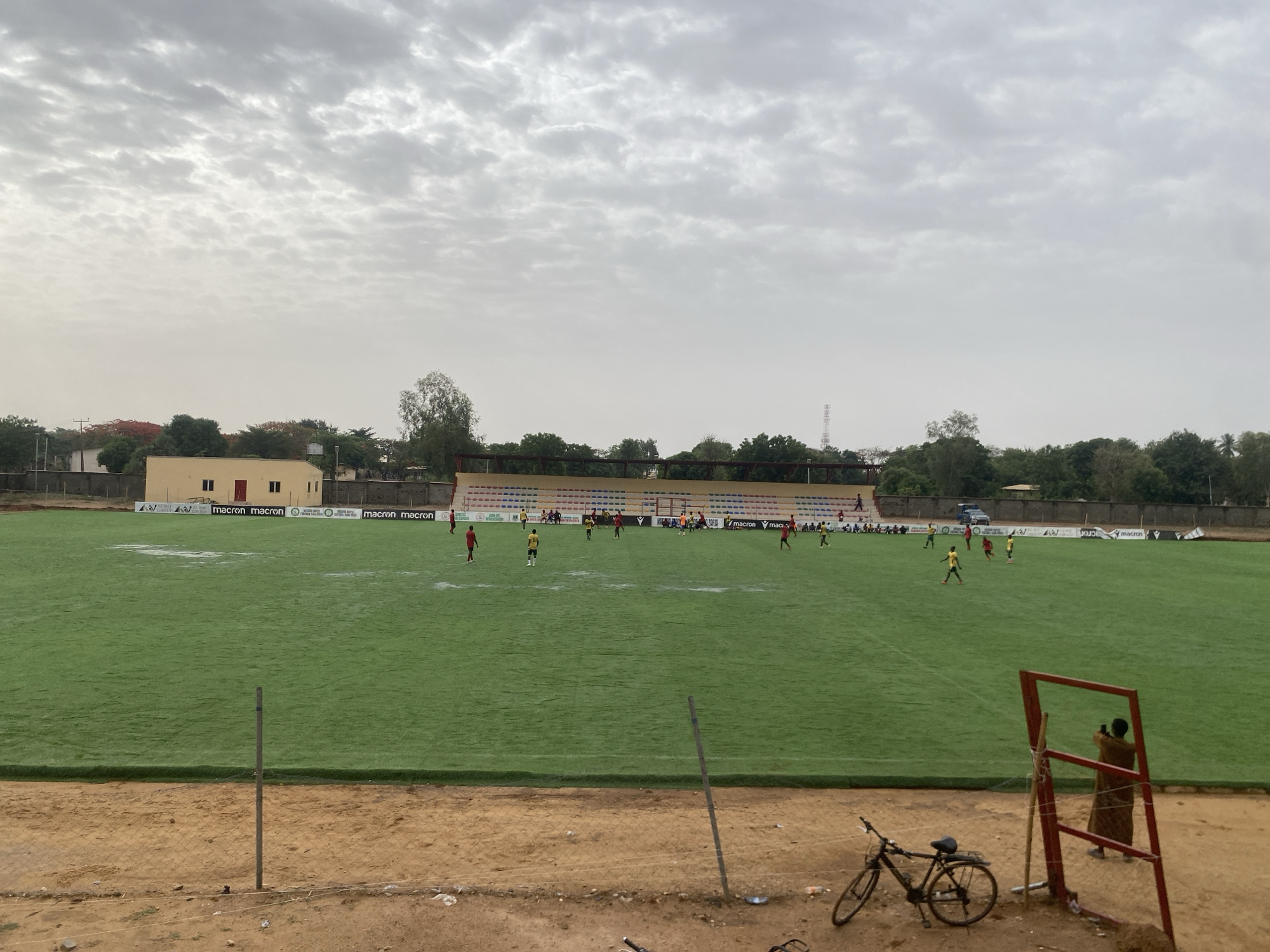 The image shows a soccer field in Tudun Wada, Nigeria, with a game in progress. Several players wearing red and yellow jerseys are spread across the field. A small bleacher section is visible behind one goal. In the foreground, there's a bicycle leaning against the edge of the field and a man in traditional clothing taking a picture. The environment appears dusty, with overcast skies. A building with yellow walls is situated at one edge of the field. The field is lined with Macron banners.