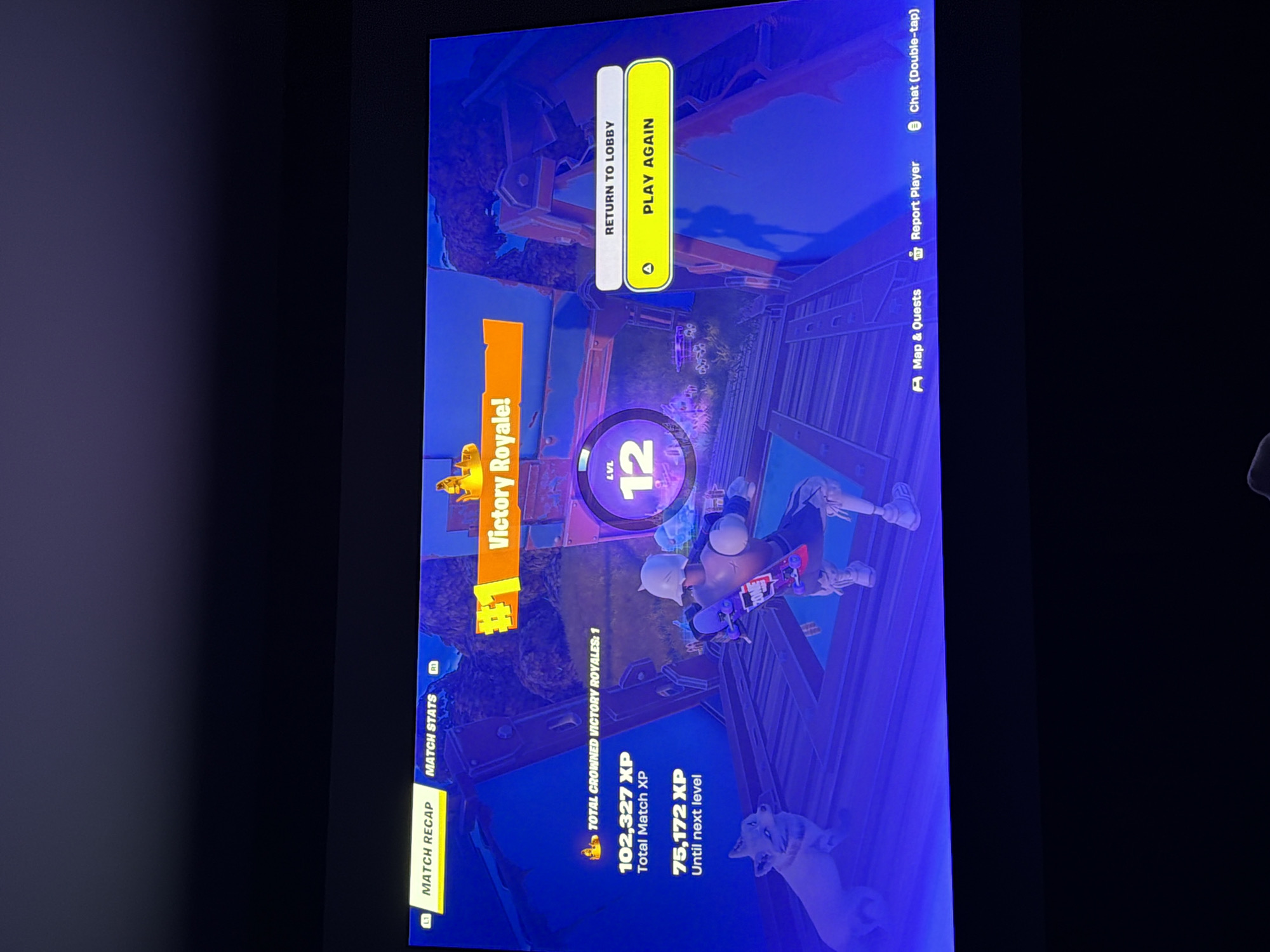 The image shows a television screen displaying a victory screen from the video game Fortnite. The on-screen text indicates a "Victory Royale!" and the player's level is 12. The player has a total of 102,327 XP and needs 75,172 XP to reach the next level. The screen offers options to "Return to Lobby" or "Play Again." Icons for "Map & Quests," "Report Player," and "Chat" are visible along the right side of the screen.