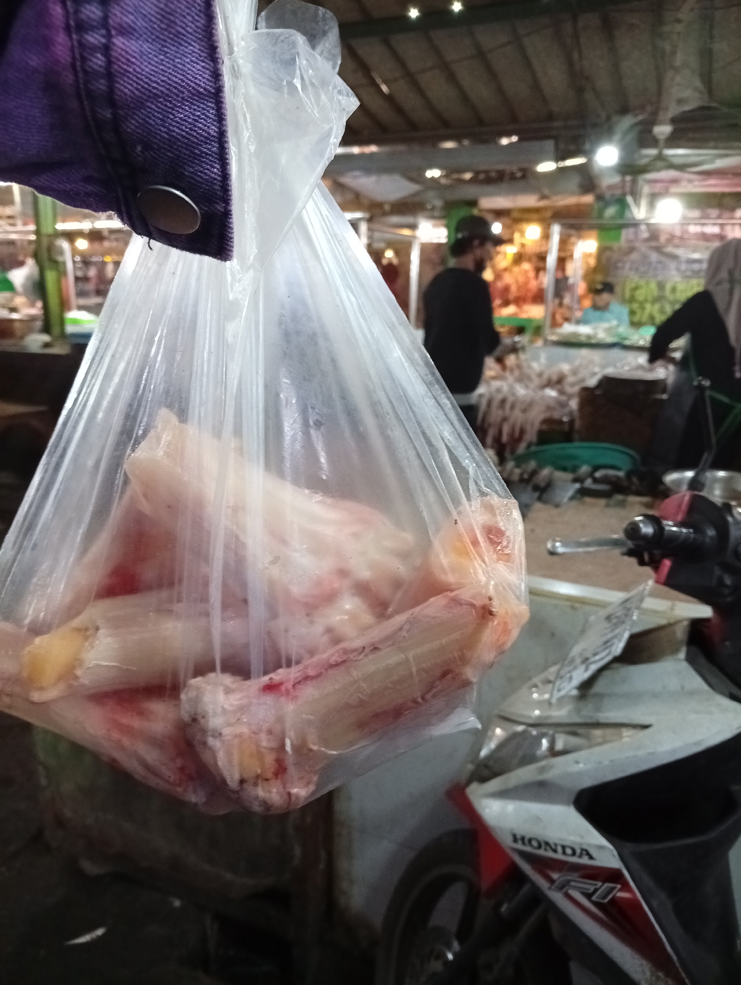 A plastic bag filled with raw meat, likely beef bone marrow, hangs in the foreground, obscuring a market scene. The bag is held by a purple garment with a metal snap. In the background, a bustling marketplace is visible, with stalls and people. One man in a dark shirt and hat is seen at a stall. To the right, a red and white motorcycle is partially visible, with the brand "HONDA" clearly displayed. The lighting suggests it is daytime, possibly indoors or under a covered area. The overall impression is a scene of commerce and food preparation in a market setting.