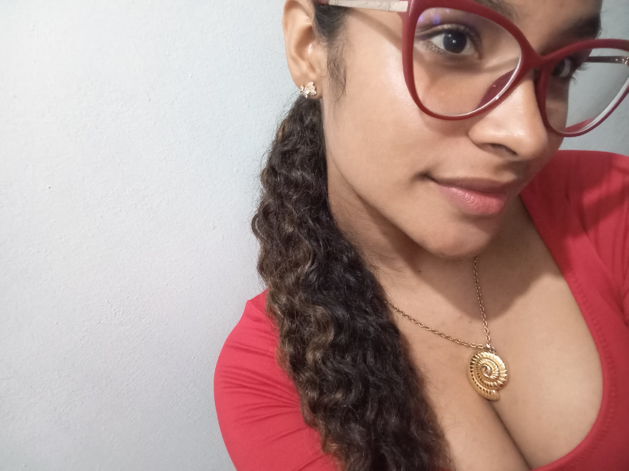 A close-up, chest-up portrait features a person, likely a young woman, partially visible against a plain white wall. She is looking towards the viewer with a subtle, soft smile, her eyes visible behind red-framed cat-eye style eyeglasses. Her dark, curly hair is styled to her right, falling over her shoulder and extending down her back.
She wears a small, intricate gold-colored stud earring in her visible right ear, which appears to have a floral or butterfly design. Around her neck is a gold-colored chain necklace with a prominent, spiraled, shell-like pendant resting on her chest. She is dressed in a vibrant red top with a softly draped, low neckline.
The background is a simple, unadorned white wall with a slightly textured surface. The lighting is even, suggesting an indoor setting or diffused natural light. The overall mood is relaxed and pleasant. There is no visible text in the image. This photograph was taken in Juan Griego, Venezuela.