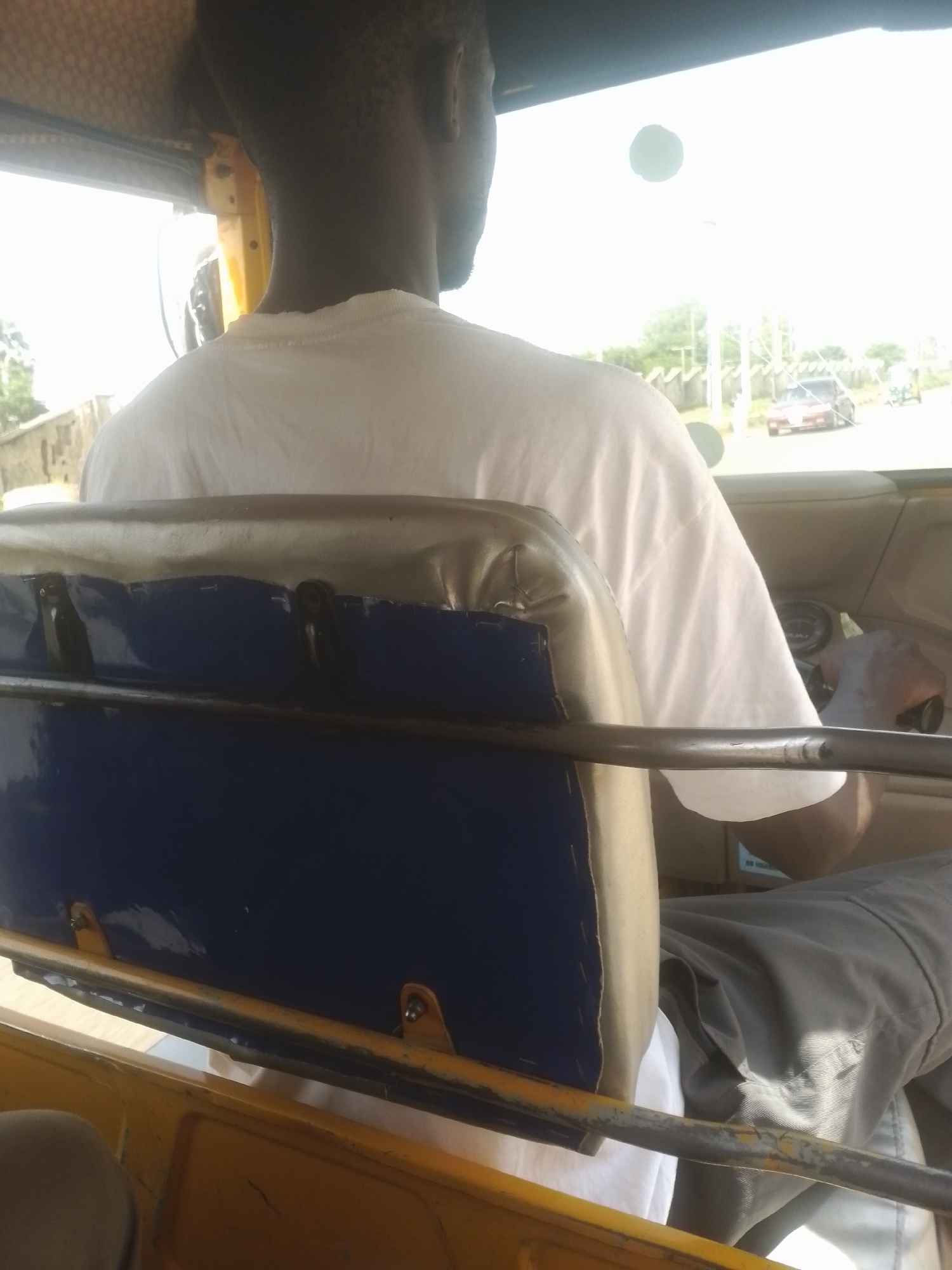 The media file shows the interior of a three-wheeled vehicle, likely a tuk-tuk, from the perspective of a passenger sitting behind the driver. The driver, a person with dark skin and short hair, is wearing a white t-shirt and is seen from the back and side. They are operating the vehicle, with their right hand visible on the steering wheel and their left hand resting on their lap.
The foreground is dominated by the back of the passenger seat, which is upholstered in blue fabric and has a padded top in a light tan color. A metal frame runs across the top of the seat. The interior of the vehicle appears to be yellow.
Through the windshield, a bright, overcast sky is visible. In the distance, there is a road with a red car driving on it, and further back, some trees and buildings can be seen. A utility pole with lights is also present. The overall lighting suggests it is daytime, possibly late morning or afternoon. There are no obvious signs of specific weather conditions other than the bright sky.
The scene depicts the act of driving or riding in a motorized rickshaw. The focus is on the driver and the immediate interior of the vehicle. The image is shot from a low angle, looking forward. There is no visible text in the image.