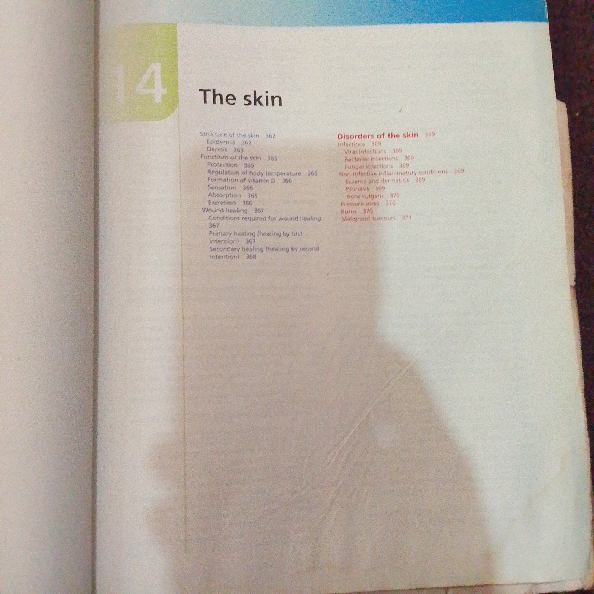 The image displays a page from a book or document. On the left side, a large number "14" is visible, printed in a light green box. To the right of the number "14" is the bolded title "The skin". Below the title, there are two columns of text, likely a table of contents or an index. The left column lists topics related to the skin such as "Structure of the skin", "Epidermis", "Dermis", "Functions of the skin", "Protection", "Regulation of body temperature", "Formation of vitamin D", "Sensation", "Absorption", "Excretion", and "Wound healing" with corresponding page numbers. The right column is titled "Disorders of the skin" and lists topics like "infections", "Viral infections", "Bacterial infections", "Fungal infections", "Non-infective inflammatory conditions", "Eczema and dermatitis", "Psoriasis", "Acne vulgaris", "Pressure sores", "Burns", and "Malignant tumours", also with page numbers. The text is printed in black and red ink. The background of the page is off-white, and the top portion of the page has a blue band across it. The image appears to be a photograph of an open book, with the right edge of the page slightly curled. There is no information in the image that would allow for the identification of its location in Jalingo, Nigeria.
