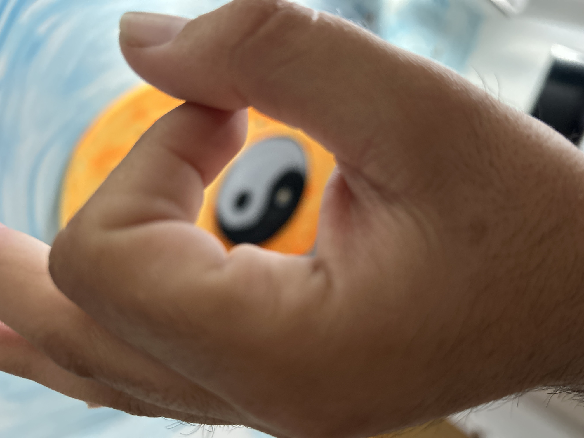 A close-up, blurred shot focuses on a hand performing the Gyan Mudra gesture, with the thumb and index finger touching to form a circle. The background is out of focus, but suggests an orange circular object with a yin-yang symbol in the center, against a light blue and white patterned surface. The setting is not identifiable as Plovdiv, Bulgaria.