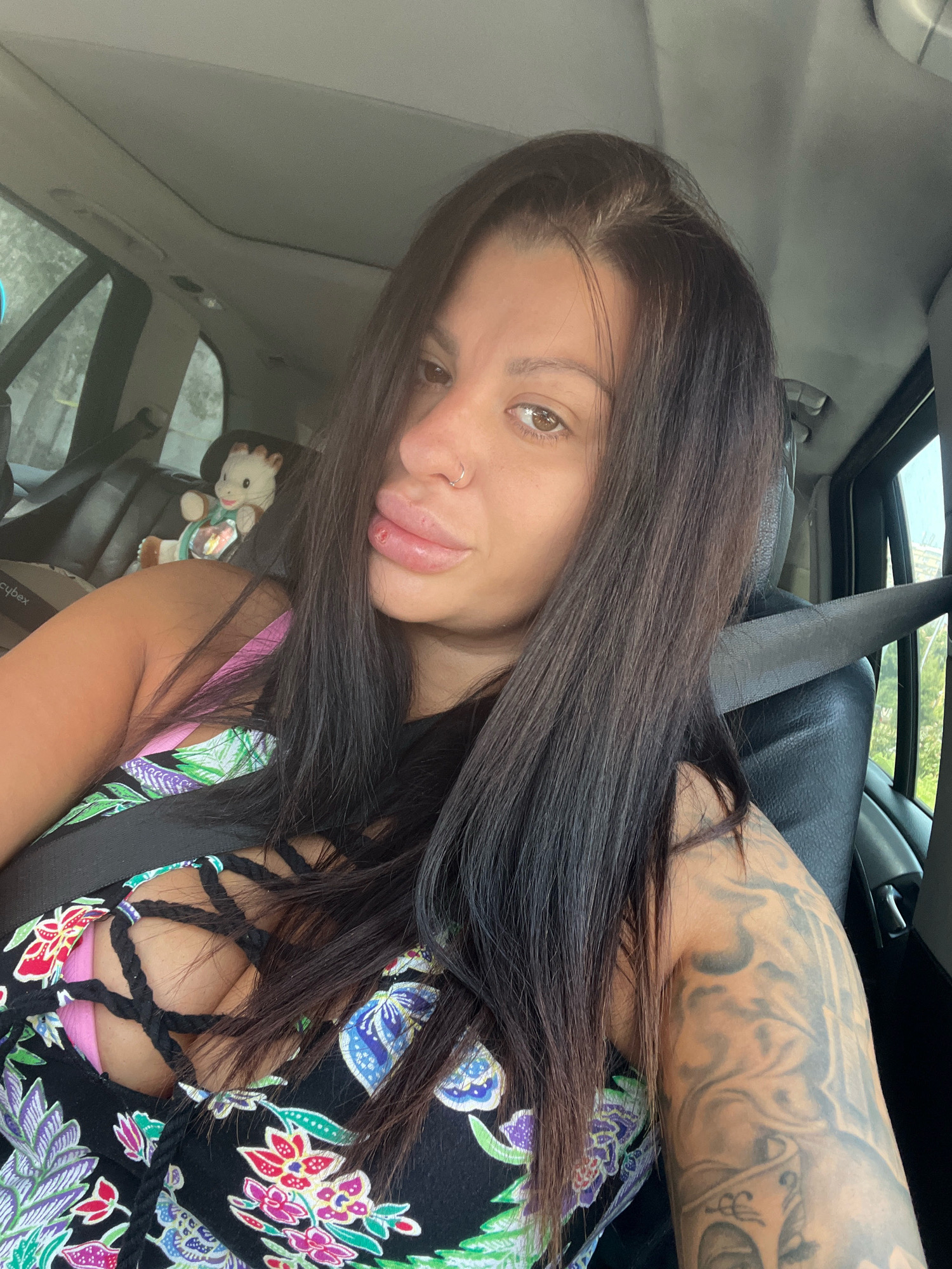 The image shows a woman sitting in the passenger seat of a car, looking towards the camera. She has long, dark hair and is wearing a black dress with a floral pattern. She has tattoos on her arm. In the background, a stuffed giraffe toy is visible in a car seat. The car appears to be parked on a street with trees and buildings visible through the windows, suggesting an urban or suburban setting. There is no specific information in the image to indicate the location is the city of Plovdiv, Bulgaria.