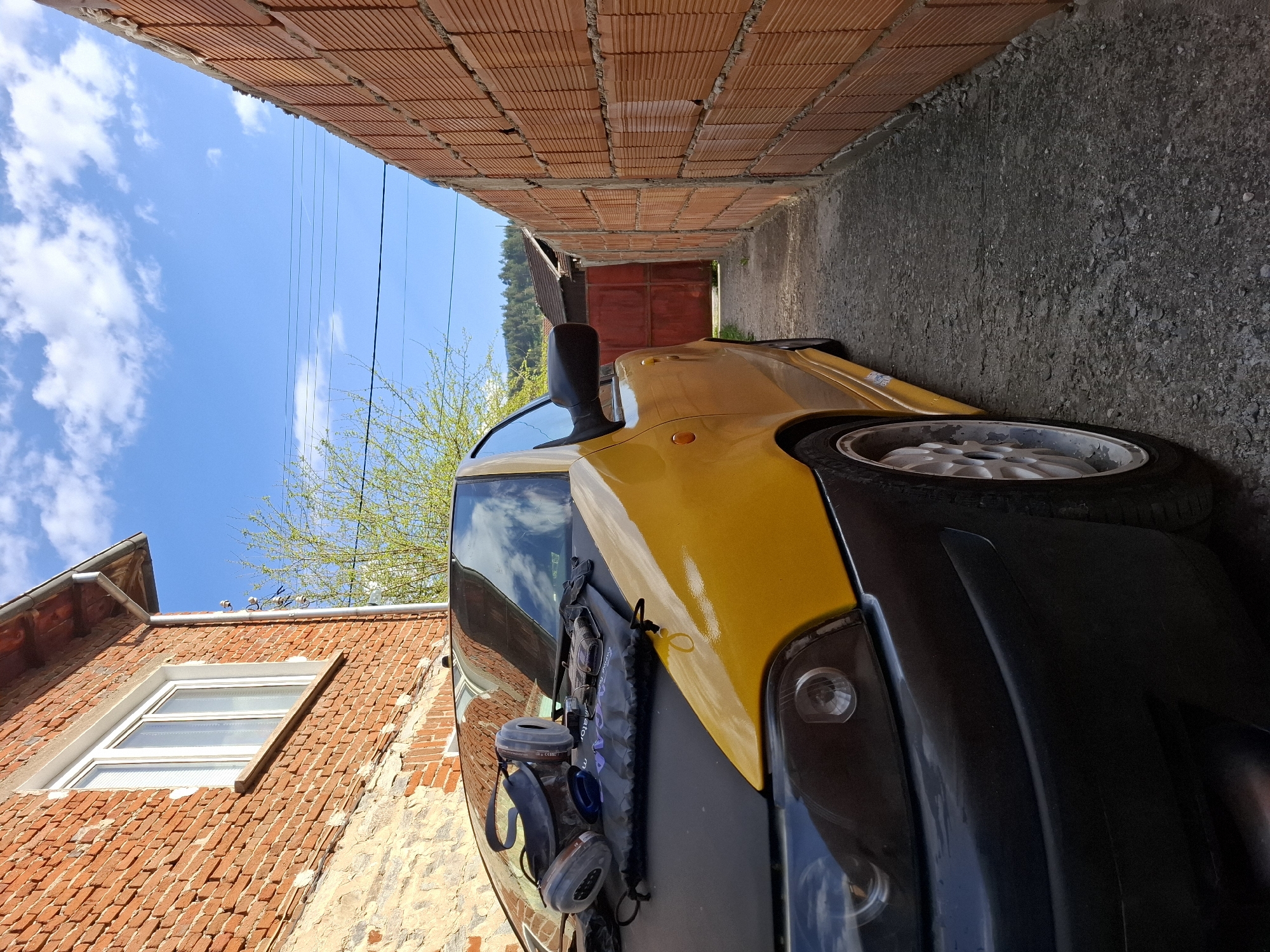 A yellow vehicle is parked tightly between two brick buildings in Rakitovo, Bulgaria. The sky is visible above, with some white clouds. The building on the left has a window. The vehicle has a black front bumper and safety gear attached to the hood.