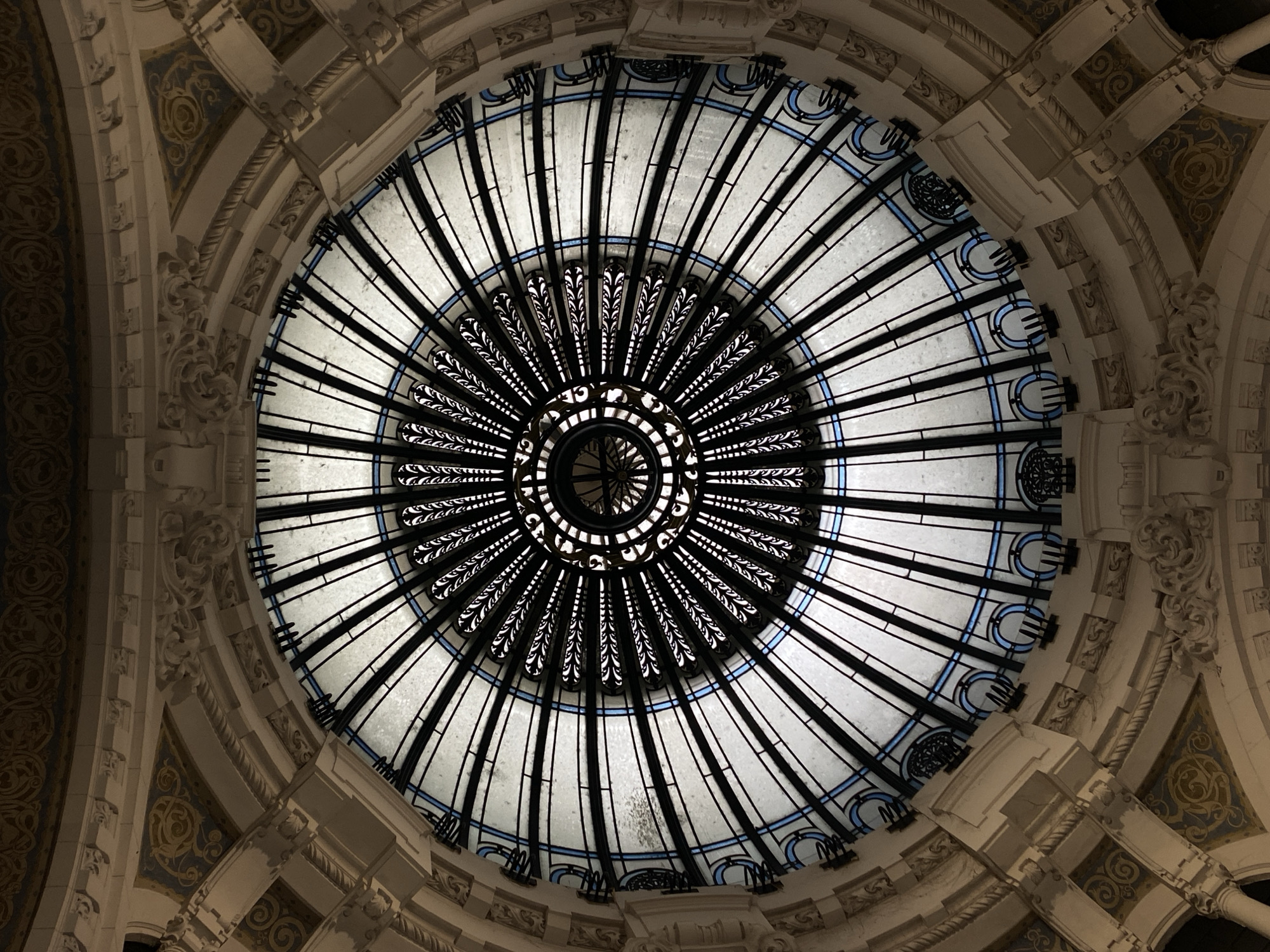 This is a photograph of the stained-glass dome in Galerias Pacifico, a shopping mall located on Florida Street in Buenos Aires, Argentina. The dome is a circular structure with intricate metalwork and translucent panels. The design is radial, with elements emanating from a central point. The metalwork consists of a series of vertical lines that radiate outward, some embellished with leaf-like patterns near the center. The panels between the metal lines appear to be made of frosted glass, with blue accents around the periphery. The dome is surrounded by ornate architecture featuring moldings, carvings, and decorative patterns. The surrounding surfaces are primarily beige, cream, and gold.