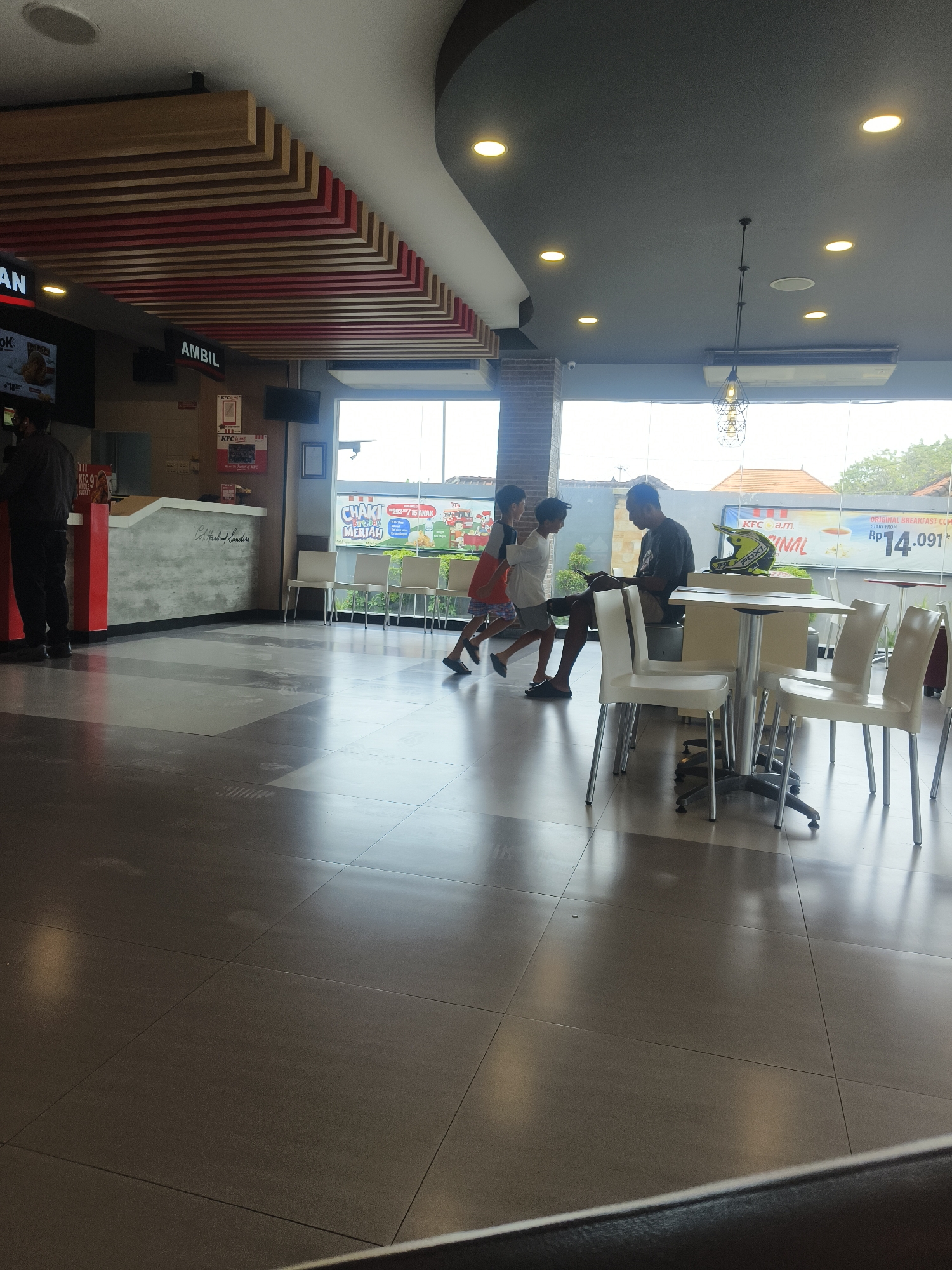 The image captures the interior of a KFC restaurant in Denpasar, Indonesia. The scene is dimly lit with fluorescent lights overhead and natural light filtering in from large windows. In the foreground, a grey tiled floor reflects the ambient light.
On the left, a man in a dark shirt and pants stands at a counter. Behind him, a red and grey counter displays a menu board with Indonesian text, including "OK" and "AMBIL" which means "take" in Indonesian.
In the middle ground, a group of people are gathered near a table. Two young boys, one in an orange shirt and shorts, the other in a white t-shirt and shorts, are walking towards a man seated at a table. The seated man wears a dark t-shirt and shorts and appears to be looking at his phone. A yellow and black motorcycle helmet rests on the table.
In the background, white chairs are arranged around tables. A large window reveals the exterior of the restaurant, with trees and buildings visible, suggesting a street-level location. A large sign with Indonesian text and cartoon characters is visible through the window. The overall atmosphere appears casual and busy.