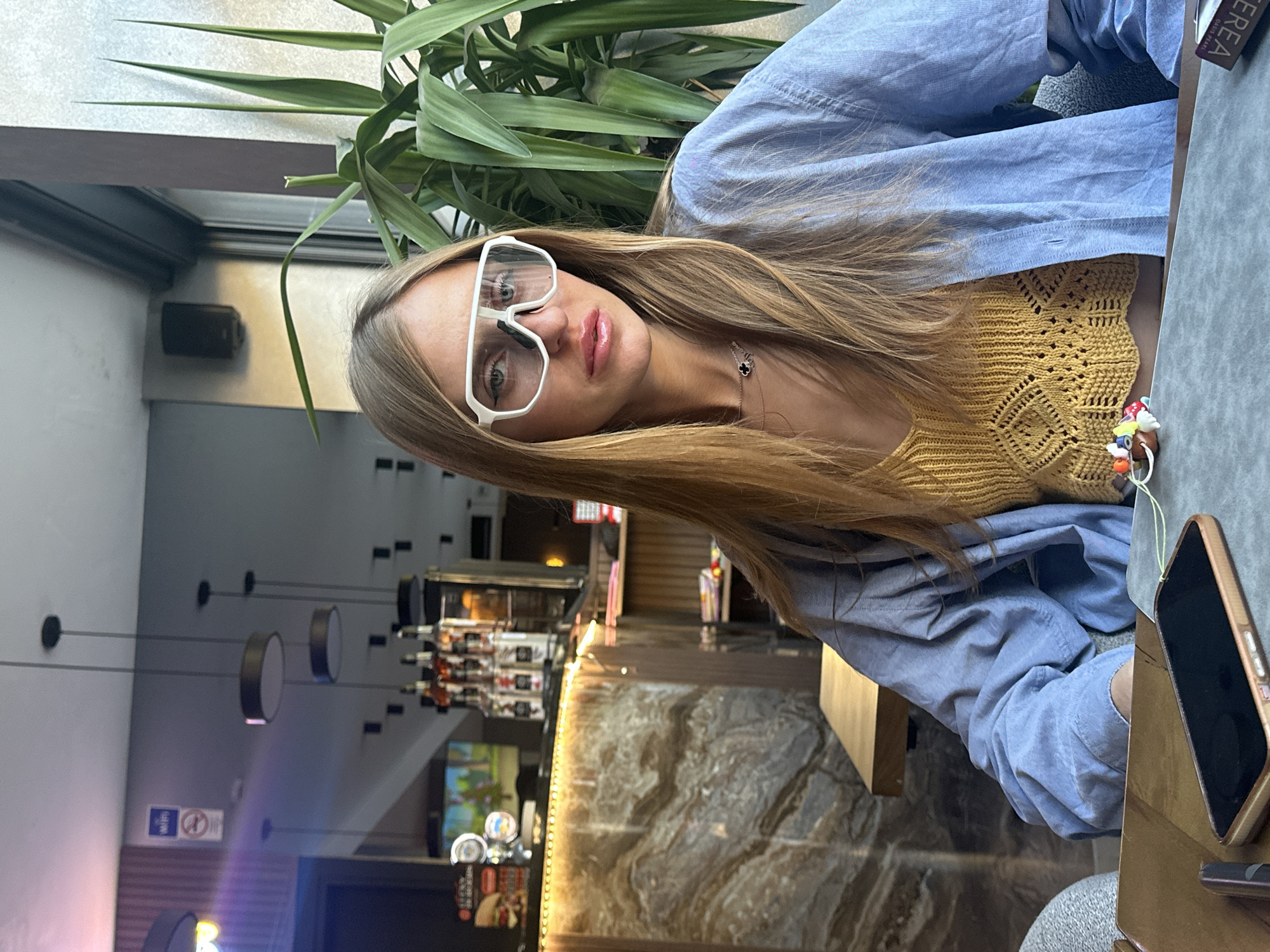 This image, taken in Budva, Montenegro, depicts a young adult female seated at a table within a cafe or bar environment. The woman, possessing long, straight, light brown hair, looks directly forward. She wears large, clear, white-framed glasses, and her lips, coated with a light pink product, are slightly parted. Her attire consists of a mustard yellow, crocheted crop top layered under an unbuttoned light blue long-sleeved shirt. A delicate gold-colored necklace featuring a black four-leaf clover charm adorns her neck.
In the immediate foreground, a dark grey table supports a smartphone in a rose gold case, placed face down. Adjacent to the phone is a cluster of colorful miniature charms strung together with a light green cord. A partially visible dark object on the table bears the text "EREA."
The background on the left features a bar area with a marble-patterned brown and tan counter, illuminated by warm LED strip lighting. Behind the counter, various bottles are arranged, and a television screen displays an animated scene. Signs indicating "WIFI" and "No Smoking" are visible on the wall above the bar, and dark, circular pendant lights hang from the ceiling. On the right side of the frame, a large green plant with spiky leaves is positioned next to a window, providing natural light. A dark speaker is mounted on the wall near the window.