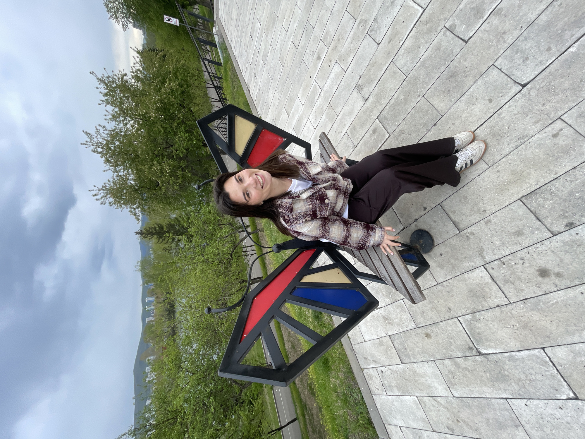 A young woman sits on a public bench, designed to resemble a butterfly's wings, on a paved area in Krasnoyarsk, Russia. She is wearing a plaid shirt, a white t-shirt, dark trousers, and white sneakers. The bench is black with sections of red, blue, and yellow. The surrounding environment includes greenery, trees, a walkway, and overcast skies. The city skyline and distant hills are visible in the background.