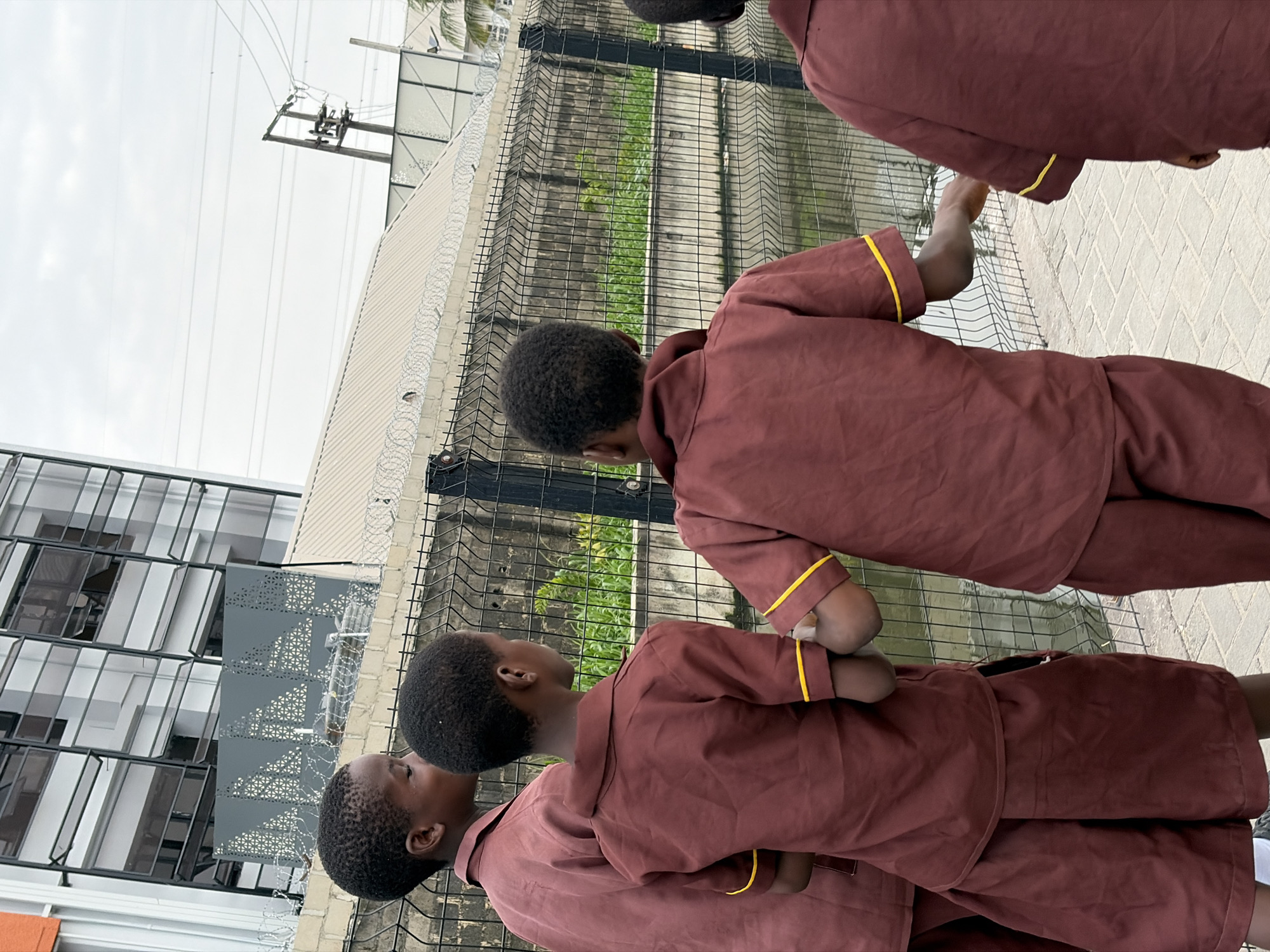 In Igboefon, Nigeria, four young boys in maroon school uniforms with yellow trim are standing by a black metal fence. The boys are facing away from the camera, looking at a body of water and green vegetation beyond the fence. One boy in the foreground has his left arm extended. Behind the fence, a light-colored building with multiple windows is visible, along with power lines and a utility pole. The ground in front of the fence is paved with light-colored bricks. The sky is overcast.