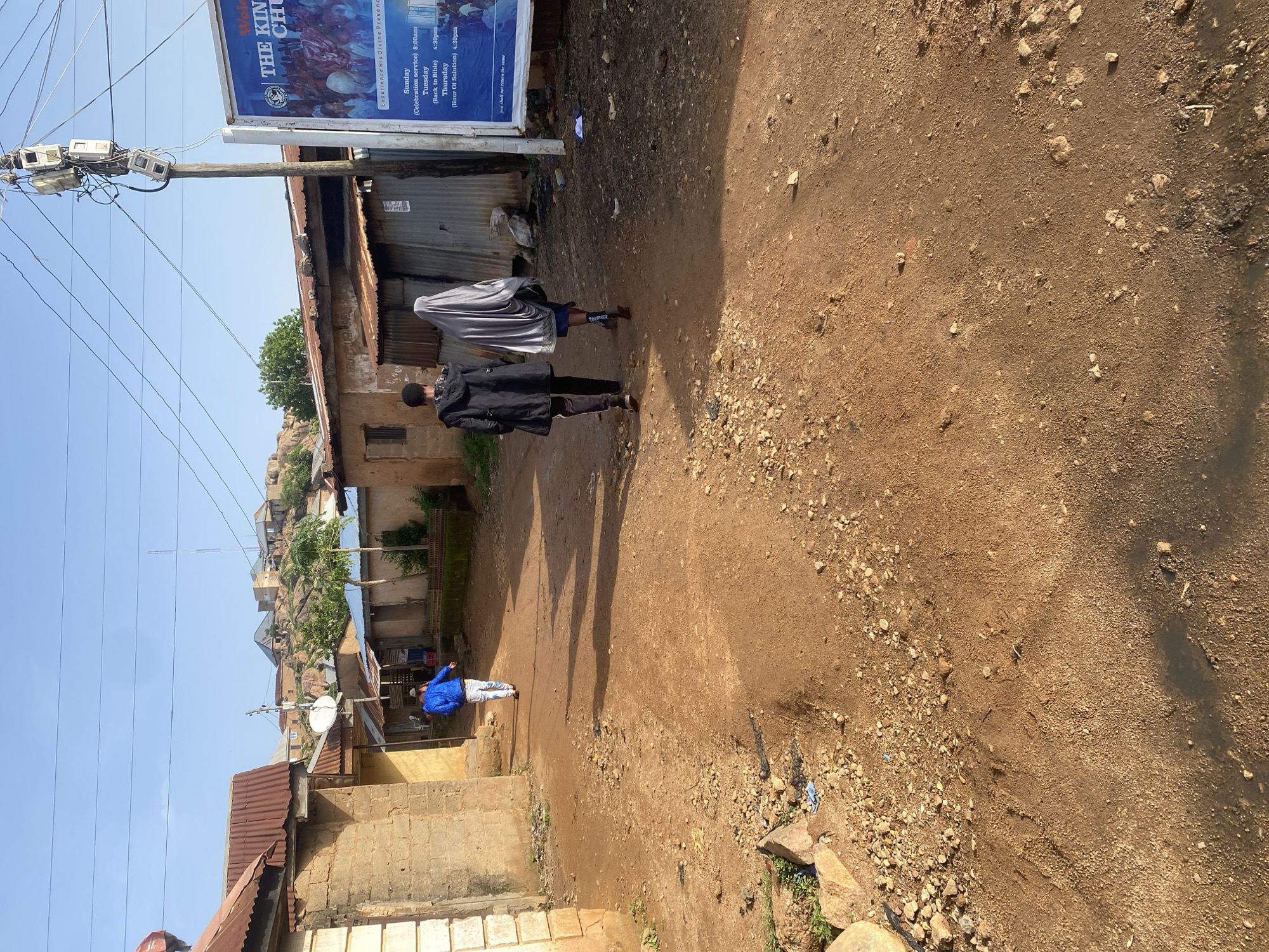 This media file captures a street scene in Jos, Nigeria, under a clear blue sky, suggesting it is daytime with fair weather. The image is oriented vertically, with the top of the scene appearing on the left.
The foreground features a dirt road, characterized by its loose soil and scattered pebbles. To the left, there are several residential structures, some with corrugated iron roofs and others with what appear to be brick walls. Electrical poles with numerous meters and tangled wires are prominent, indicating active infrastructure. A large billboard is also visible on the left, displaying text related to "THE KINGDOM CHURCH" with service times for Sunday, Tuesday, and Thursday.
The midground shows two individuals walking away from the viewer down the dirt road. One person is wearing a black jacket and pants, while the other is partially obscured by a gray or silver garment, possibly a hijab or a shawl. Further down the road, another person in a bright blue jacket and light-colored pants is visible. The shadows cast by the individuals and the structures suggest the sun is positioned to the right, likely mid-morning or late afternoon.
The background reveals more buildings and houses climbing a rocky hillside, a common topographical feature in Jos. The overall impression is of a bustling, informal urban environment, possibly a residential area with local businesses or community centers. The presence of people engaged in daily activities like walking down the street suggests normalcy. The text on the billboard provides specific information about a local church, hinting at the community's religious life.
