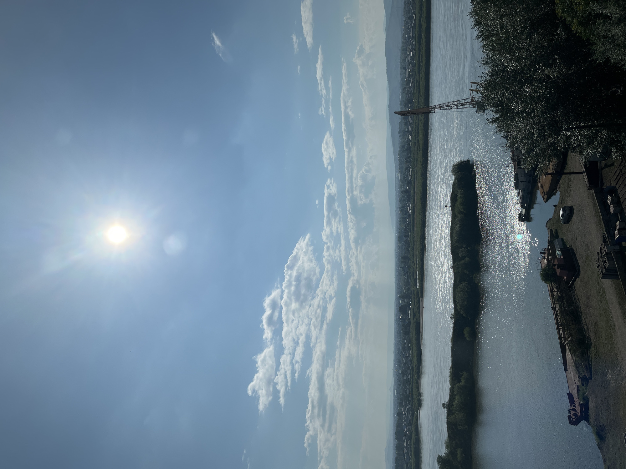 The image depicts a wide river with a small island in the middle, reflecting the bright sun. On the far bank of the river, a sprawling town with houses and trees can be seen, bordered by rolling hills in the distance. In the foreground, on the right side of the image, is a dense cluster of trees. To the left, on the riverbank, there is a barge and a crane, suggesting industrial activity. The sky is a clear blue with scattered white clouds, and the sun is prominently visible, casting a strong glare. The overall scene is one of a riverside landscape with elements of both nature and human development.