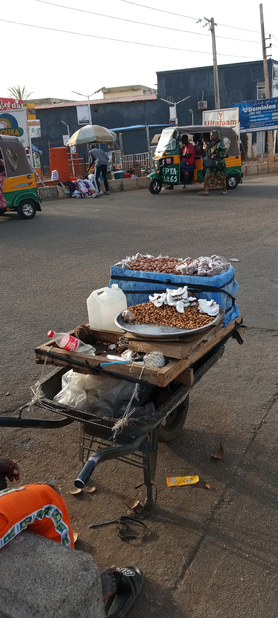The image depicts a lively street scene in Jos, Nigeria, possibly during morning or late afternoon due to the harsh shadows. In the foreground, a mobile vendor's cart is laden with goods. On a silver platter, a substantial pile of small, round, light-brown nuts (likely tiger nuts) is displayed next to several pieces of cracked coconut. Behind this, various packaged items, possibly snacks or small goods, are stacked on a blue covering. A transparent jerrycan filled with liquid and an empty red Coca-Cola bottle are also on the cart.
The mid-ground features a paved road with human activity and vehicles. A green and yellow auto-rickshaw, commonly known as a "keke napep," with the registration "PTA 16265," is parked on the road, carrying passengers. A woman in a patterned dress stands beside it. Another keke napep is partially visible on the left. On the sidewalk to the left, a person bends over a display of clothes or goods under a small, light-colored umbrella. Buildings line the background, and utility poles with overhead wires are also present.
Visible text on signs includes "Bridal," "MAX," and "QUKA." A prominent blue billboard in the background reads "Wake up & Dominion JOS HQ Every Sunday Every Wednesday," with "Oitafoam" also visible on a sign nearby. The lower left corner of the frame shows a person's legs and feet, clad in black slippers, suggesting someone is seated close to the vendor's cart, possibly interacting with the vendor. The scene conveys the busy atmosphere of a street market or commercial area.