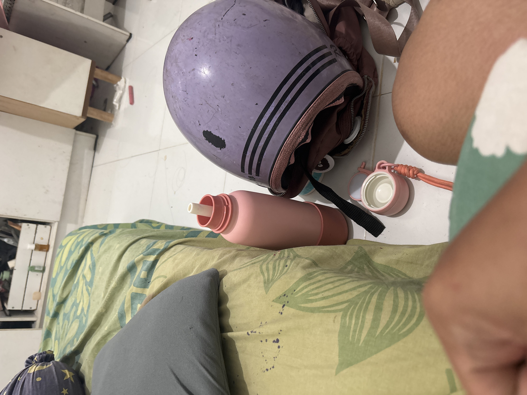 The image shows a domestic scene, likely a bedroom, with a bed covered in a yellow and green patterned comforter. A gray pillow rests on the comforter. On the floor beside the bed are several items: a light pink water bottle with a white spout, a purple helmet with black stripes resting on a brown bag, and a small pink container with an orange strap. A person's leg and arm are visible in the foreground. In the background, part of a white cabinet with wooden accents is visible, and on a shelf within the cabinet, there are several small bottles and containers. The flooring appears to be tiled. The scene is consistent with a personal living space. There is no specific visual information to indicate a location in North Jakarta, Indonesia.