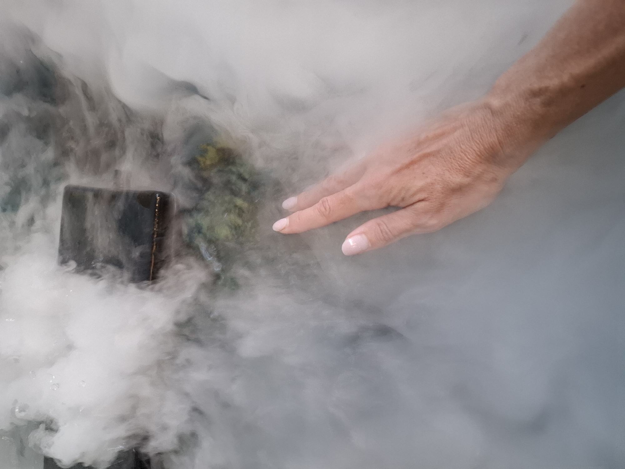A person's hand with light pink nail polish is submerged in thick, white fog, obscuring the lower arm. The fog obscures a dark, rectangular object and a blurry, green, and yellow textured surface beneath. The scene is likely an indoor environment given the contained fog and the presence of a submerged object. No specific location in Nha Trang Ward, Vietnam, can be identified from the visual information.