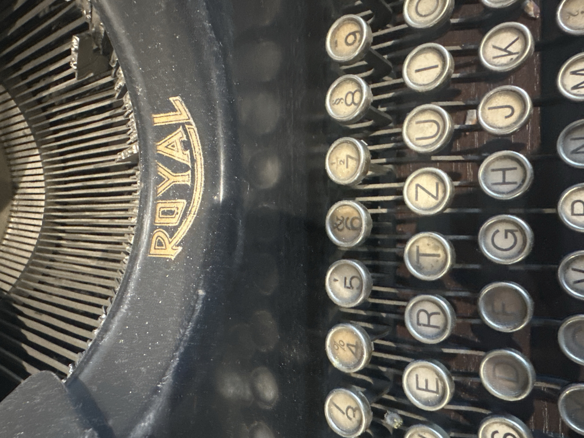 This image is a close-up, eye-level shot of the keyboard and typing mechanism of an antique Royal typewriter. The typewriter is black with gold lettering that reads "ROYAL" across the top. The keys are arranged in rows, with some keys displaying numbers and symbols, and others displaying letters of the alphabet. The typing hammers, which strike the ink ribbon to imprint characters on paper, are visible behind the keys. The scene appears to be indoors, with the lighting suggesting an ambient, possibly natural light source. There is no indication of the city of Łódź, Poland in the image.