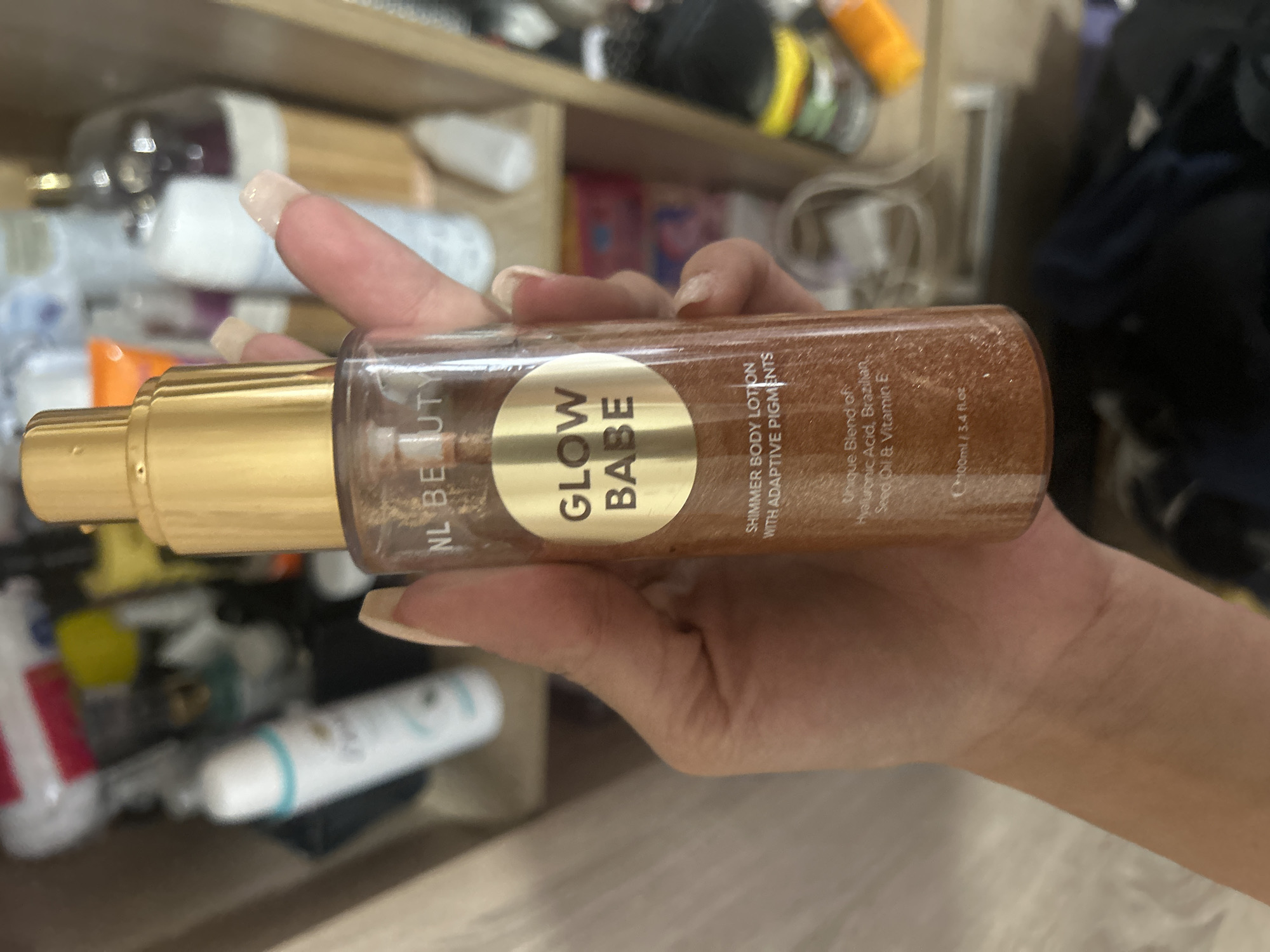 A hand with manicured nails holds a bottle of "GLOW BABE" shimmer body lotion. The bottle has a gold pump and is filled with a shimmering bronzed lotion. The label on the bottle reads "NL BEAUTY" at the top and below that, a gold circle contains the words "GLOW BABE." Below the circle, it states "SHIMMER BODY LOTION WITH ADAPTIVE PIGMENTS" and then "Unique Blend of Hyaluronic Acid, Brazilian Seed Oil & Vitamin E". At the bottom of the bottle, it indicates "100ml / 3.4 fl.oz". The background appears to be a shelf with various cosmetic products and other items, suggesting a personal collection or storage area. The image is not geographically specific to Perushtitsa, Bulgaria, nor does it provide any information that would place it in that location.