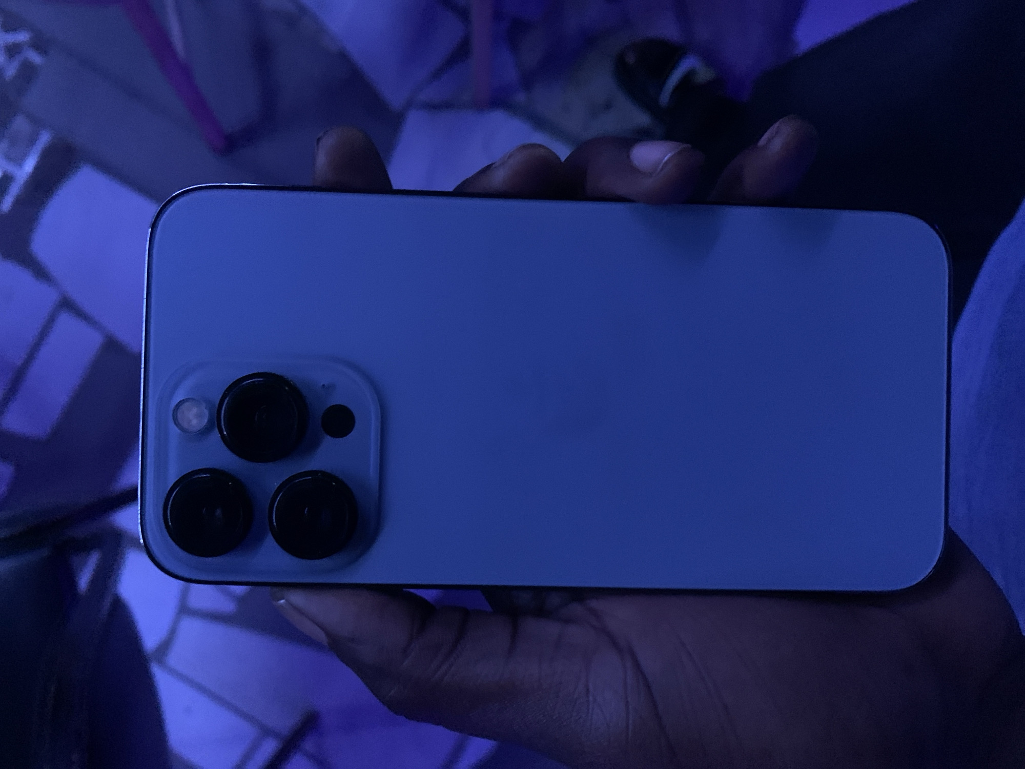 A blue iPhone is held in someone's hand. The camera lens array on the back of the phone is visible. The background is blurred and blue. The image does not contain enough information to determine the location in the city of New Stadium, Nigeria.