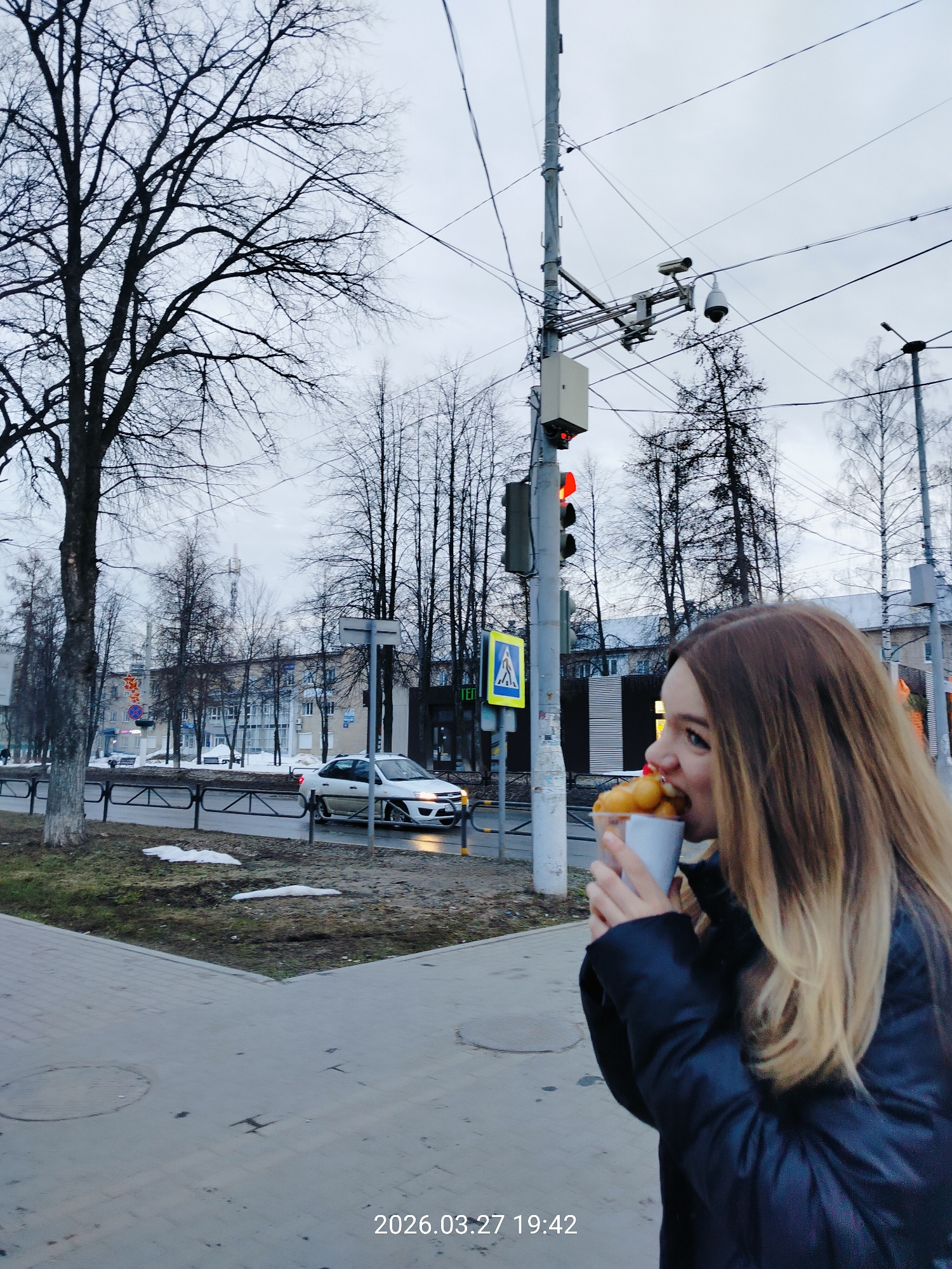 The image presents an urban street scene in Chaikovsky, Russia, captured at 19:42 on March 27, 2026, under an overcast, light grey sky. In the immediate foreground, to the right, a female with long, light brown to blonde hair and a dark jacket is shown in profile, actively eating small, golden-brown, spherical items from a white paper cup. Her mouth is open, engaging with the food. She stands on a concrete sidewalk, which exhibits a circular manhole cover. Adjacent to the sidewalk are patches of dirty snow and sparse, dormant grass.
In the midground, a black metal barrier separates the sidewalk from an asphalt road. A light-colored sedan, possibly white or silver, travels on the road from left to right, with its headlights illuminated. A tall, grey utility pole dominates the center-left, featuring a red traffic light, a blue pedestrian crossing sign with a white walking figure, and two surveillance cameras mounted on cross-arms (one a dome camera, the other a bullet-style camera). Numerous electrical wires extend from the pole and crisscross the upper frame.
The background consists of a line of bare-branched deciduous trees, indicative of late winter or early spring. Behind the trees, several multi-story buildings are visible, typical of an urban environment, some displaying illuminated signs with Cyrillic text.