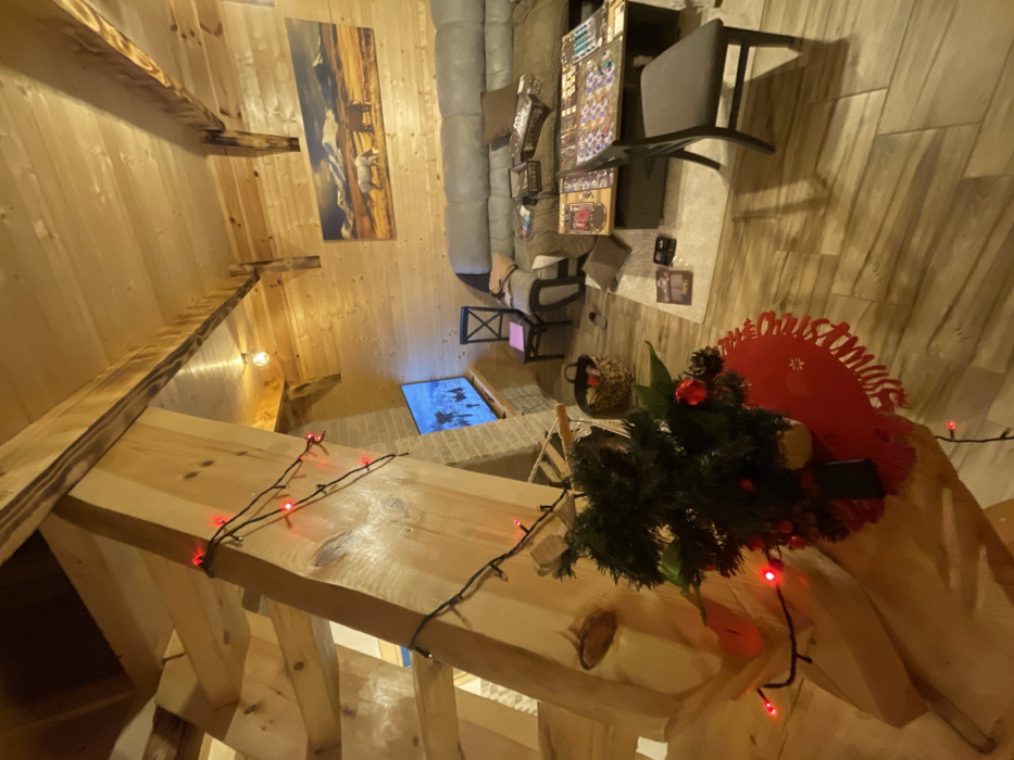 An interior view of a room in the city of Palež, Montenegro, is depicted, characterized by extensive light-toned wood paneling on the walls and ceiling, featuring exposed wooden beams. In the left foreground, a substantial wooden staircase structure, adorned with red LED string lights, rises diagonally. Positioned on a surface in front of the staircase is a small, artificial tabletop Christmas tree decorated with red ornaments and pinecones, resting on a red, scalloped placemat with a snowflake pattern. A dark object is partially visible beneath the tree.
To the right, a large, L-shaped grey sectional sofa with brown cushions is visible. In front of the sofa are two dark-colored low tables. The table closer to the sofa displays several board game boxes, including one labeled "Gloomhaven." The table further forward has a board game fully set up and in progress, featuring numerous game components. A dark grey upholstered chair is positioned at this table. To the left of the sofa, a single dark wooden chair is present, with a pink object on its seat, and a woven basket rests on the floor nearby.
On the back wall, a rectangular painting depicting two horses grazing in a field with snow-capped mountains hangs above the sofa. In the left background, a television screen, mounted on a light-colored brick structure, displays a scene of multiple figures on a snowy landscape, possibly engaged in winter sports. The room is illuminated by warm overhead lighting from an exposed bulb and other fixtures. The floor is covered with light-colored plank flooring. No individuals are visible in the scene.