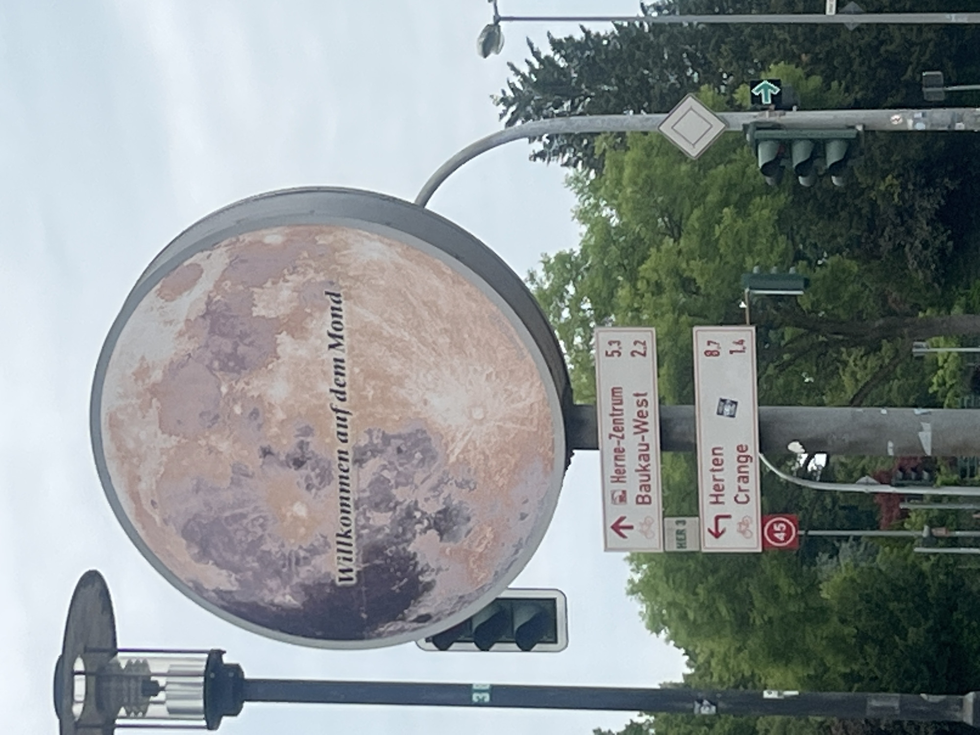 The image shows a large, circular sign depicting the moon with the text "Willkommen auf dem Mond" (Welcome to the Moon) superimposed on it. Below this sign, there are several directional street signs. One sign points upwards and to the right, indicating "Herne-Zentrum" (Herne Center) and "Baukau-West" with corresponding distances of 5.3 and 2.2 respectively. Another sign points to the left, indicating "Herten" and "Crange" with distances of 8.7 and 1.4. To the left of the directional signs, a traffic light is visible. Further down the pole, a speed limit sign with the number "45" inside a red circle is present. The environment appears to be an urban setting with trees and a cloudy sky. The presence of the directional sign for "Herne-Zentrum" confirms the location in Herne, Germany.