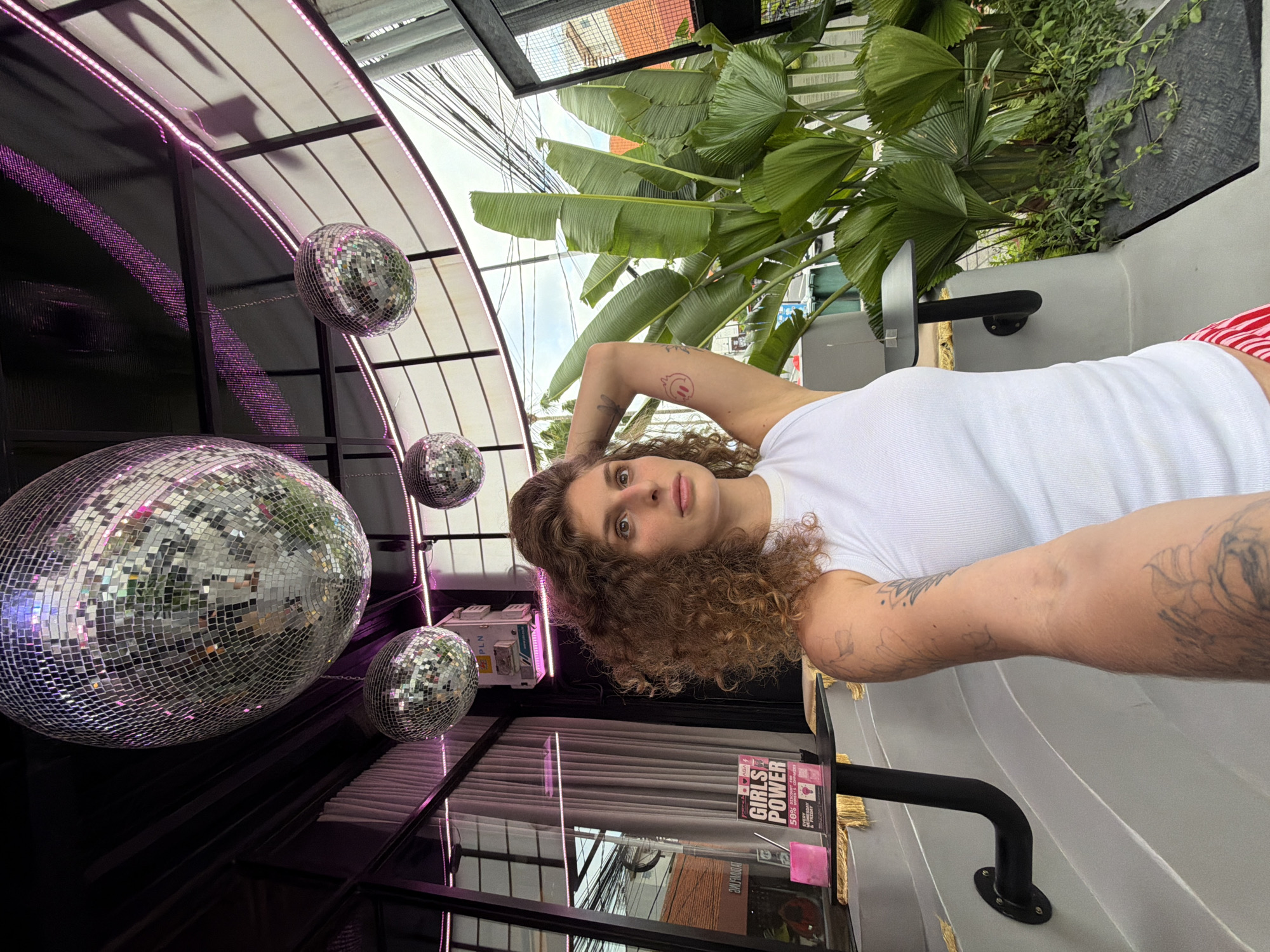 A woman with curly brown hair is seated outdoors, leaning back with her right arm raised and hand behind her head. She is wearing a white tank top and red and white striped shorts. She has tattoos on her left arm and shoulder. Behind her, lush green tropical plants are visible. Several disco balls are hanging from the ceiling above, and pink LED lights are strung along the ceiling structure. A small table with a sign that reads "GIRLS POWER" is positioned to her left. The scene appears to be an outdoor seating area of a venue, possibly a cafe or bar, in North Kuta, Indonesia, with overhead power lines and buildings visible in the background.