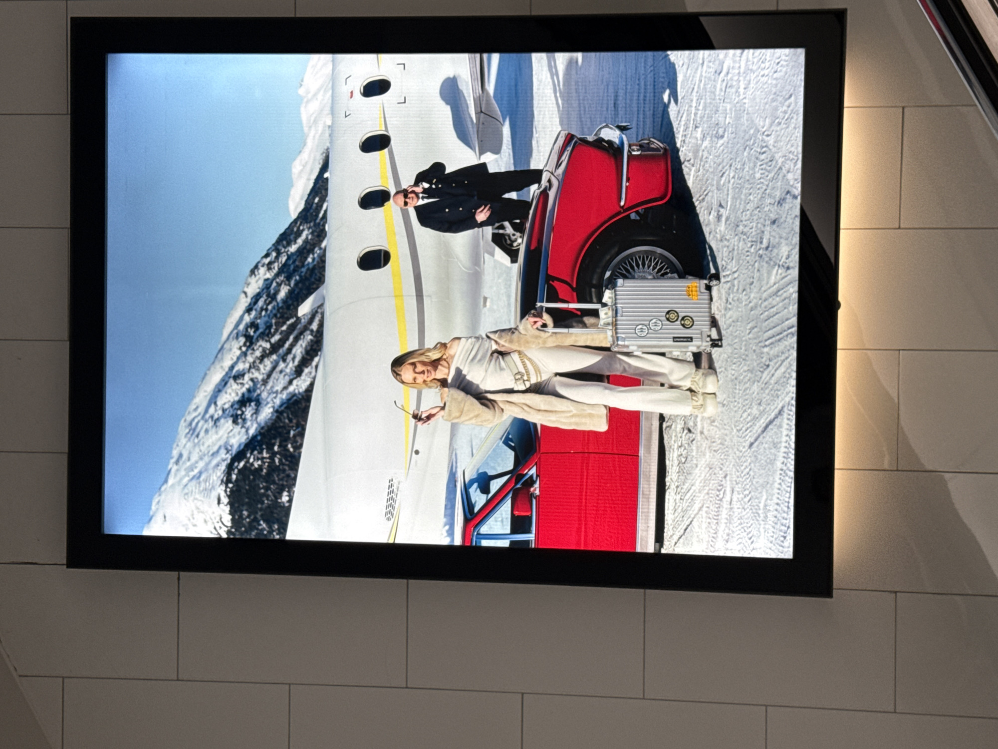 The image shows a large, illuminated advertisement displayed in what appears to be a public space in Sesto San Giovanni, Italy, possibly a shopping mall or transportation hub, given the tiled wall. The advertisement depicts a glamorous scene set against a backdrop of snow-covered mountains and a private jet. A woman in a white off-the-shoulder top and matching pants, accessorized with a fur coat and a belt bag, stands next to a bright red vintage car. She holds a pair of sunglasses in her hand. Beside her is a silver suitcase with stickers. A man in a dark suit and tie stands behind the car, appearing to be on a mobile phone. The ground is covered in snow, with tire tracks visible. The advertisement is framed by a black border and is lit from behind.