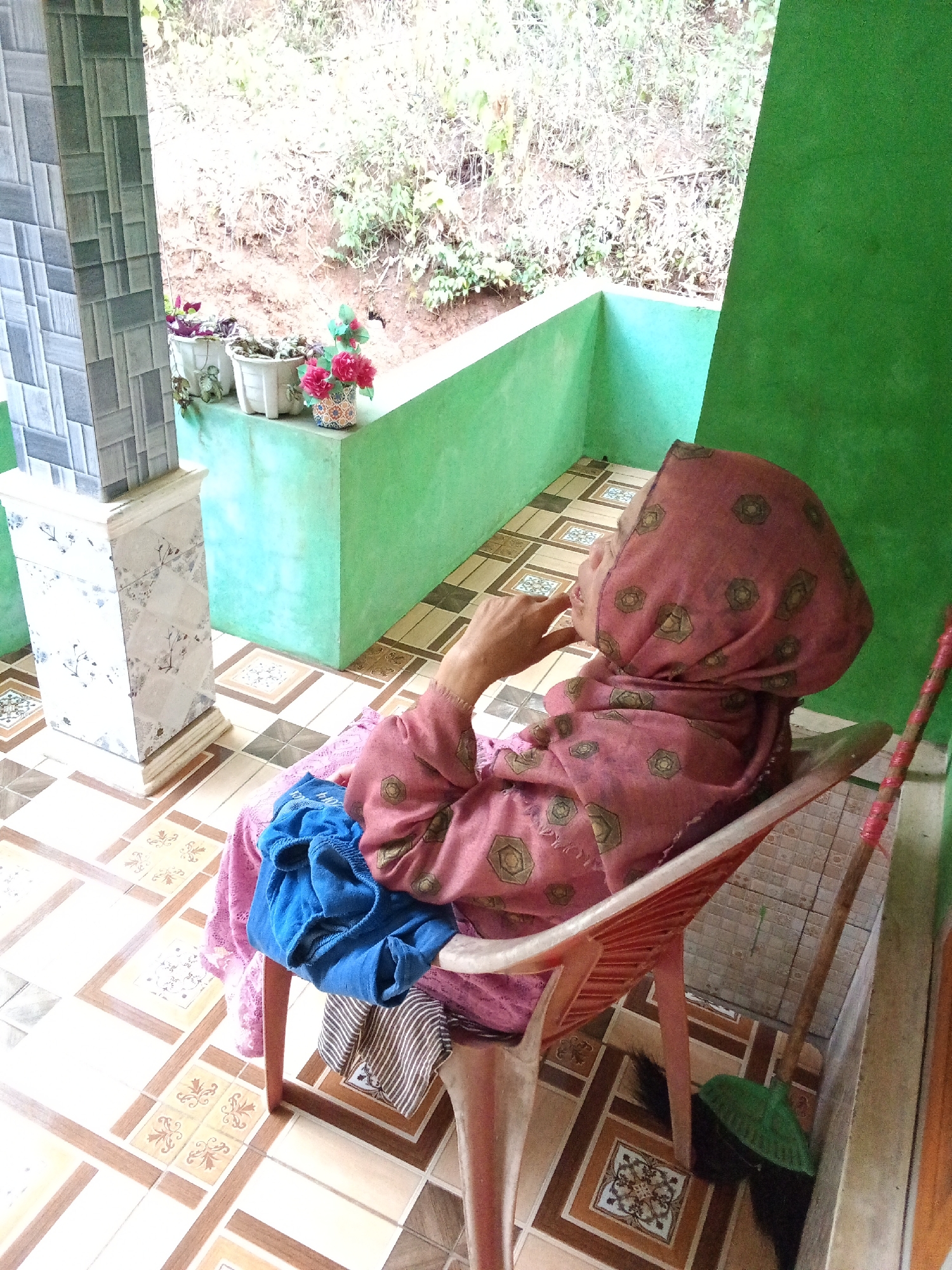A person wearing a pink hijab and a pink patterned garment is seated on a red plastic chair on a tiled porch. A blue cloth and a striped fabric are draped over the chair. A broom with a green head and a wooden handle is leaning against the wall. The porch has a green wall and is open to an outdoor area with vegetation and a slope. In the background, there are three potted plants, two with green leaves and one with pink flowers. A decorative tiled pillar is visible on the left. The floor is covered in a pattern of light and brown square tiles. The scene appears to be an outdoor seating area of a residential property in Subang, Indonesia.