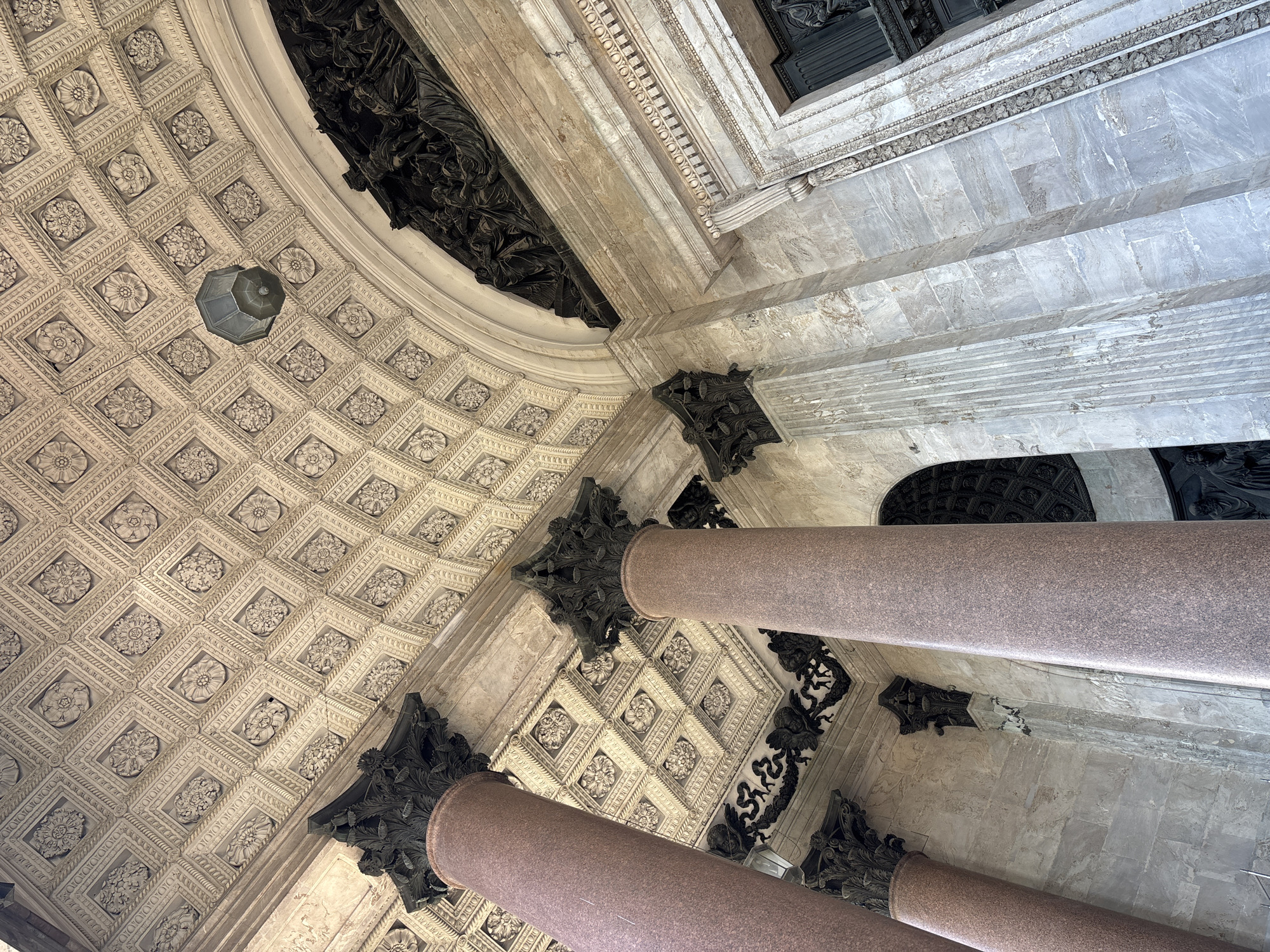 The image captures a low-angle view of the exterior of Saint Isaac's Cathedral in Saint Petersburg, Russia. Dominating the foreground are two massive pink granite columns, topped with ornate dark green capitals. Behind these columns, a richly decorated ceiling is visible, featuring a grid of square panels, each containing intricate floral motifs and decorative borders. A dark, octagonal lantern hangs from the ceiling. Above the columns and to the right, a large, arched entryway is framed by marble stonework. A dark relief sculpture is mounted above the arch. The walls are clad in light-colored marble, some sections featuring vertical fluting. The overall impression is one of grandeur and elaborate architectural detail. There are no people visible in the frame.