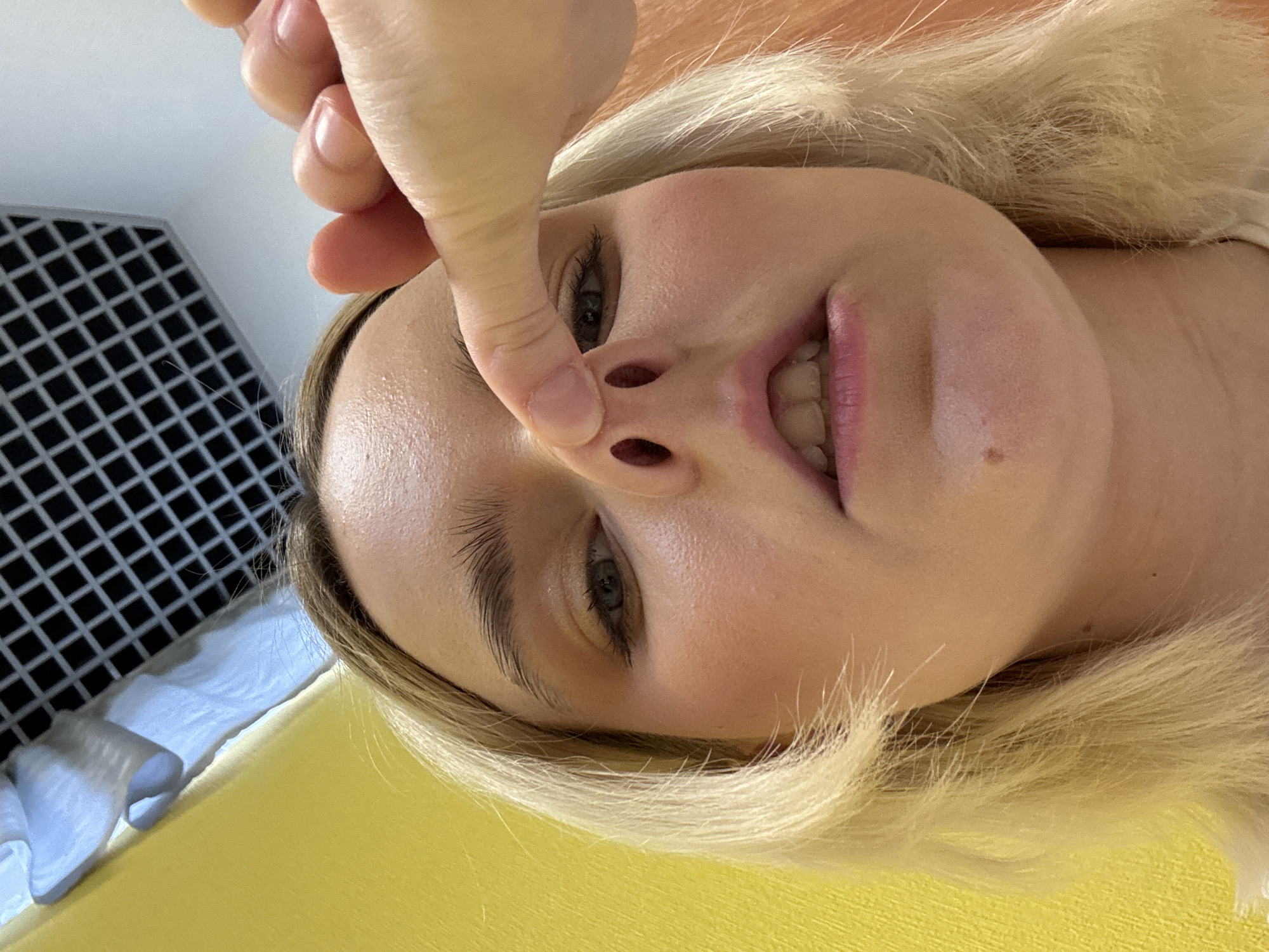 A blonde-haired woman is pictured from a low angle. A pale-skinned hand, presumably belonging to another person, holds one of her nostrils open. The woman is smiling, revealing her teeth. The background is blurred but suggests a modern indoor environment. There is no information to suggest the photo was taken in Saint Petersburg, Russia.