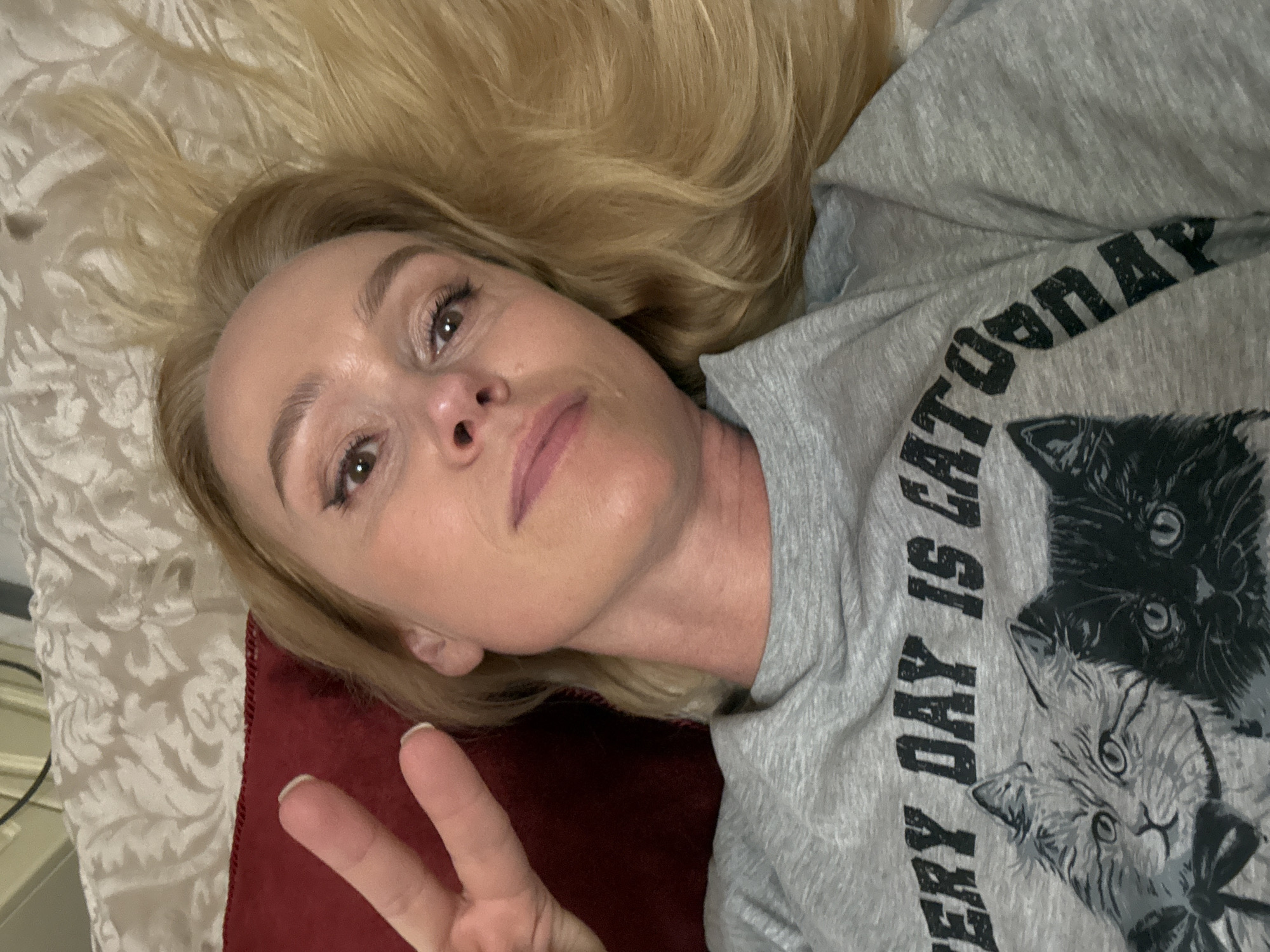A blonde woman is lying on her back on a bed with an off-white patterned comforter and a red pillow, making a peace sign with her left hand. She is wearing a gray sweatshirt with a print of cats and the text "EVERY DAY IS CATURDAY". The location of the image is unknown.