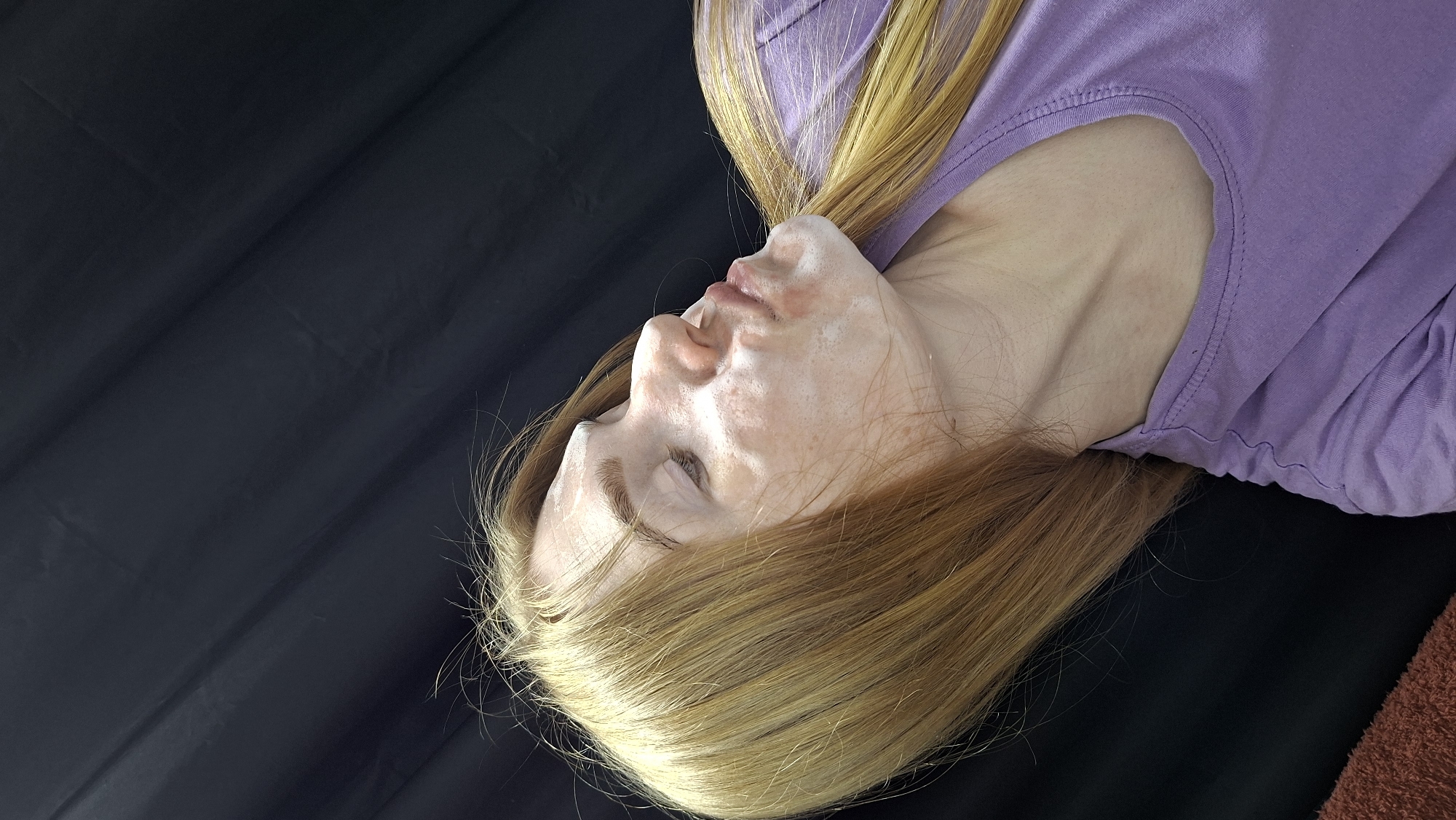 A young, fair-skinned person lies supine against a black background, wearing a light purple t-shirt. The person's face appears to have a white coating or cream applied to it. The hair is light blonde at the roots, fading into a reddish-brown towards the ends. The person's eyes are closed. There is no discernible evidence suggesting the scene is located in the city of Chaikovsky, Russia.