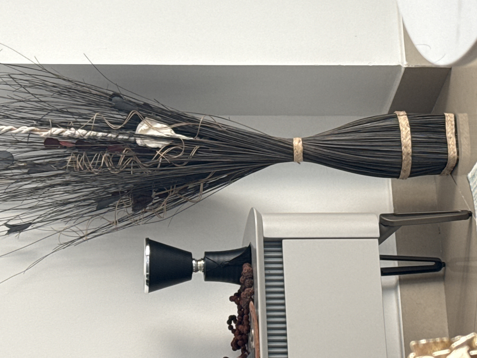 The image displays a still life composition within an indoor setting, possibly a home or a showroom in Dubai, United Arab Emirates. In the foreground, a tall, slender decorative vase crafted from dark reeds is positioned on the floor. The vase is cinched with two bands of woven material, one near the top and another near the base, giving it a bottle-like silhouette. Adjacent to the vase, on the right side of the frame, a portion of a white circular object, likely a table or stool, is visible.
To the left of the vase stands a light-colored cabinet with a ribbed front panel. On top of the cabinet, a black conical object with a silver rim rests. To the left of this object, a cluster of small, reddish-brown dried berries or fruits is scattered. The cabinet is supported by slender, dark-colored legs. The overall environment appears clean and modern, with a neutral-toned wall in the background. The lighting is soft and diffused, suggesting an indoor, possibly daylight, setting. There are no people or animals present in the image.