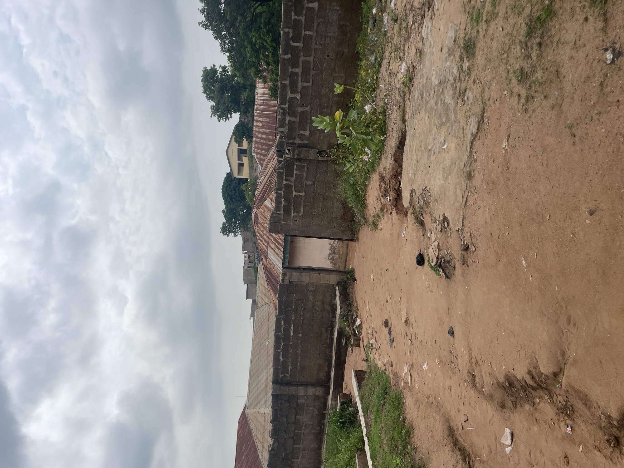 The image shows a dirt path leading towards a gate in a brick wall in Lokoja, Nigeria. The path is uneven and scattered with debris, including plastic bags and other refuse. To the left of the path is a patch of grass and some small concrete structures. The brick wall appears aged and weathered. Beyond the wall, several buildings with corrugated metal roofs are visible, along with lush green trees and a cloudy sky. The overall scene suggests a somewhat unkempt environment within a residential or semi-residential area. There are no people or animals visible in the image.