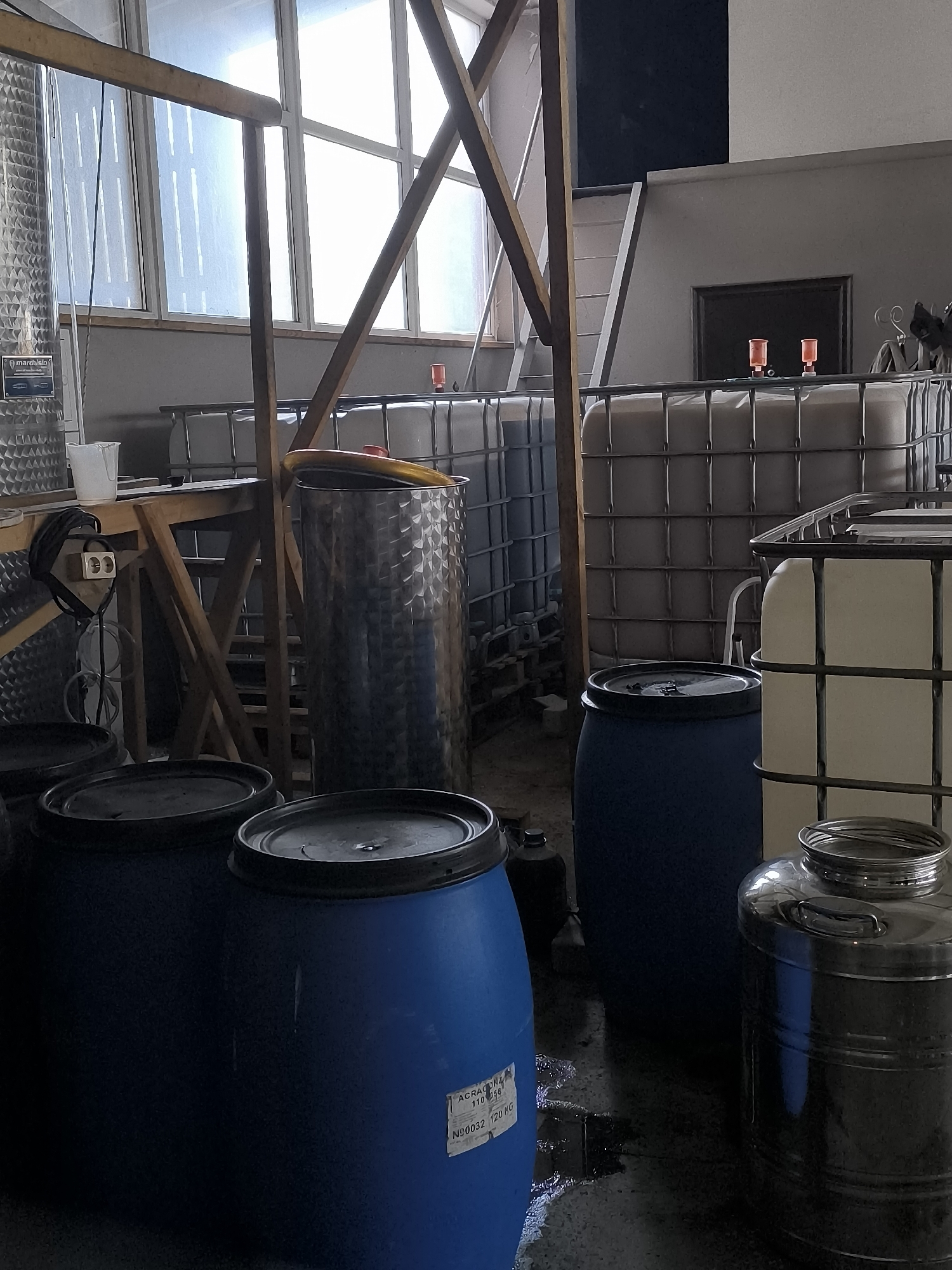 The image depicts an indoor storage area, likely a workshop or facility, filled with various containers. In the foreground, two large blue plastic barrels with black lids are visible. To the right, a stainless steel cylindrical container with a lid and handles is present. Further back, a large, patterned stainless steel drum stands upright. Behind these, several large white IBC (Intermediate Bulk Container) totes, typically used for liquid storage, are arranged in rows, some with metal cages around them. The environment appears industrial, with concrete flooring and large windows with diffused light filtering through. Wooden support beams are visible in the background, along with some electrical outlets and cables. The overall impression is one of a storage or processing space for liquids. The specific location in Kuldīga, Latvia is not determinable from the visual information provided in the image.
